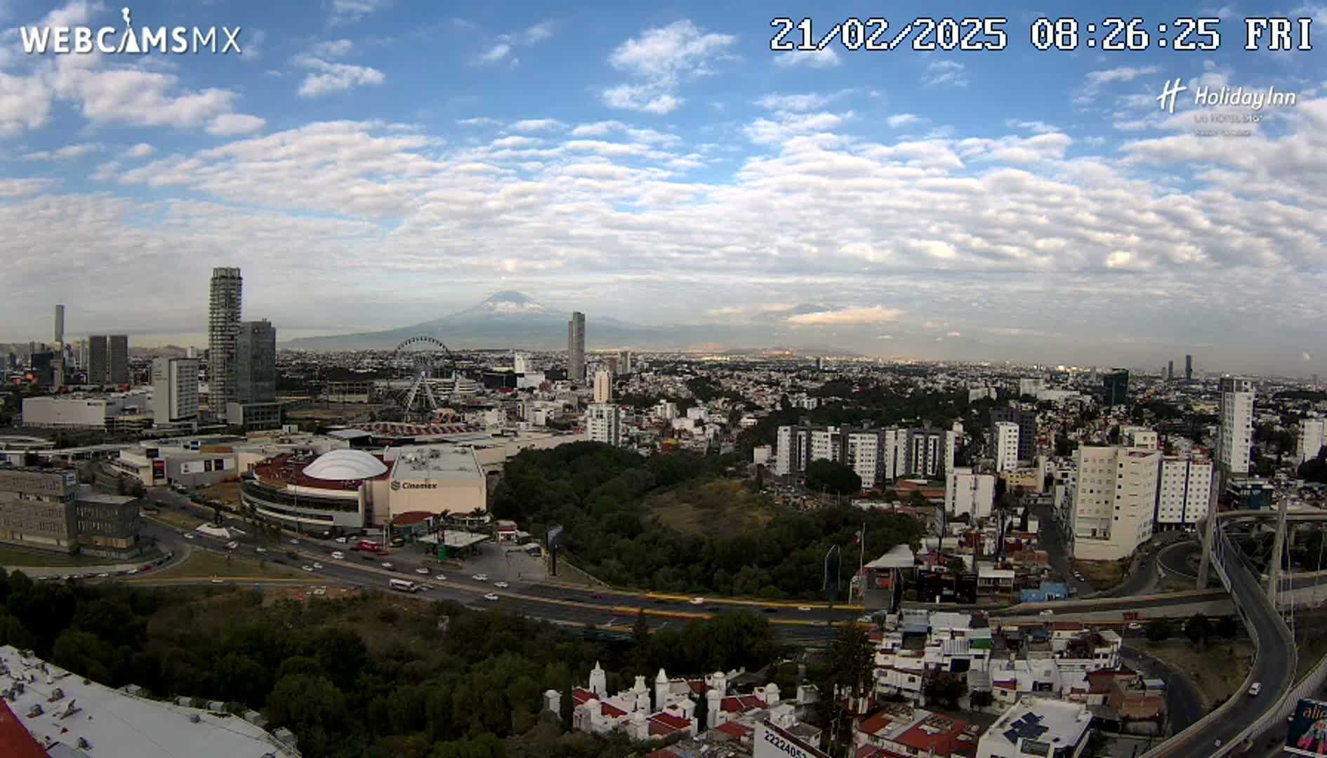 A partly cloudy day reveals a sprawling city with a Ferris wheel, numerous high-rises, a volcano in the distance, and a highway system weaving through the urban landscape.