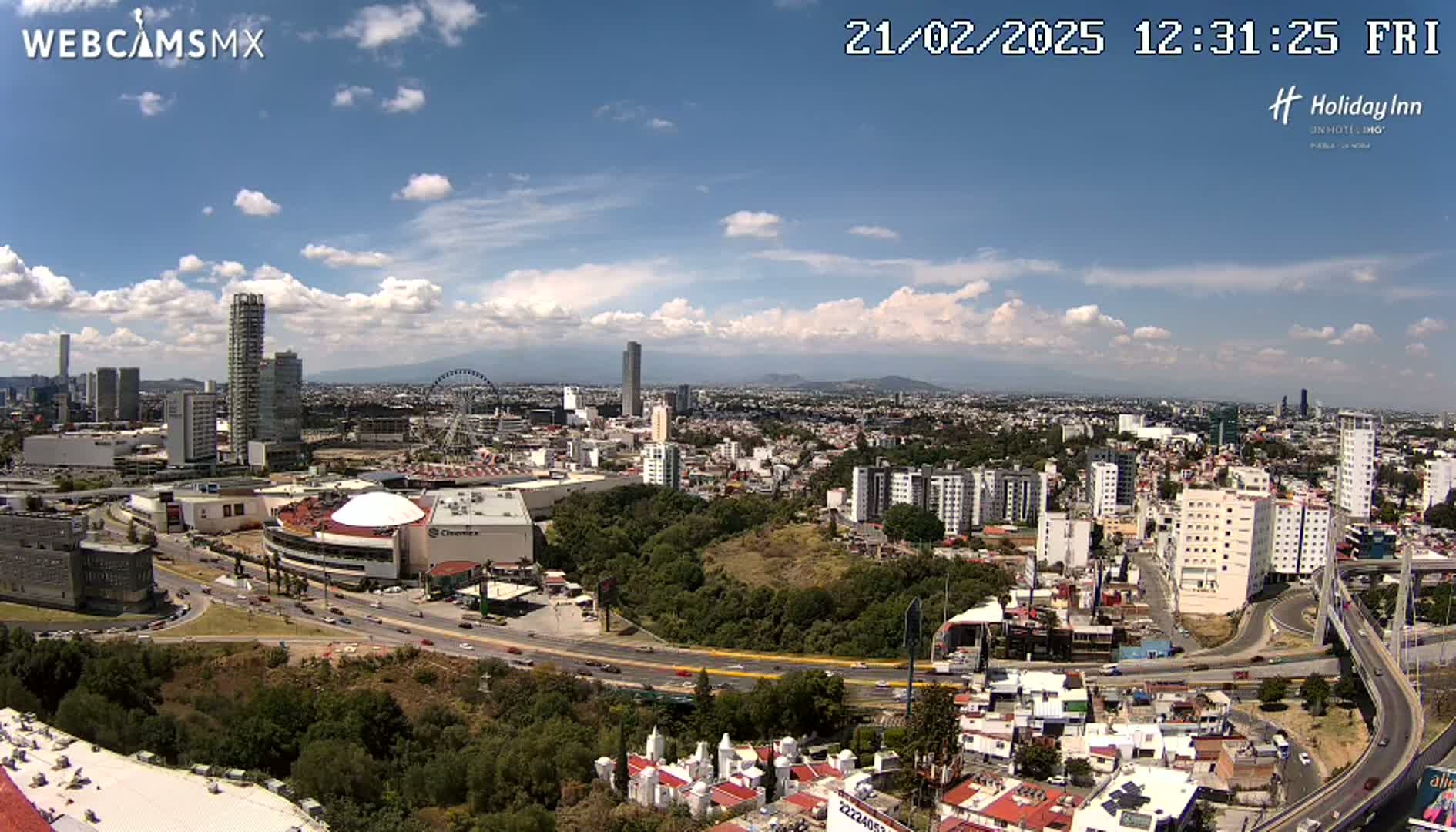 A partly cloudy day reveals a sprawling cityscape with numerous high-rise buildings, a Ferris wheel, and a large shopping mall, all situated near a major highway and surrounded by green areas.