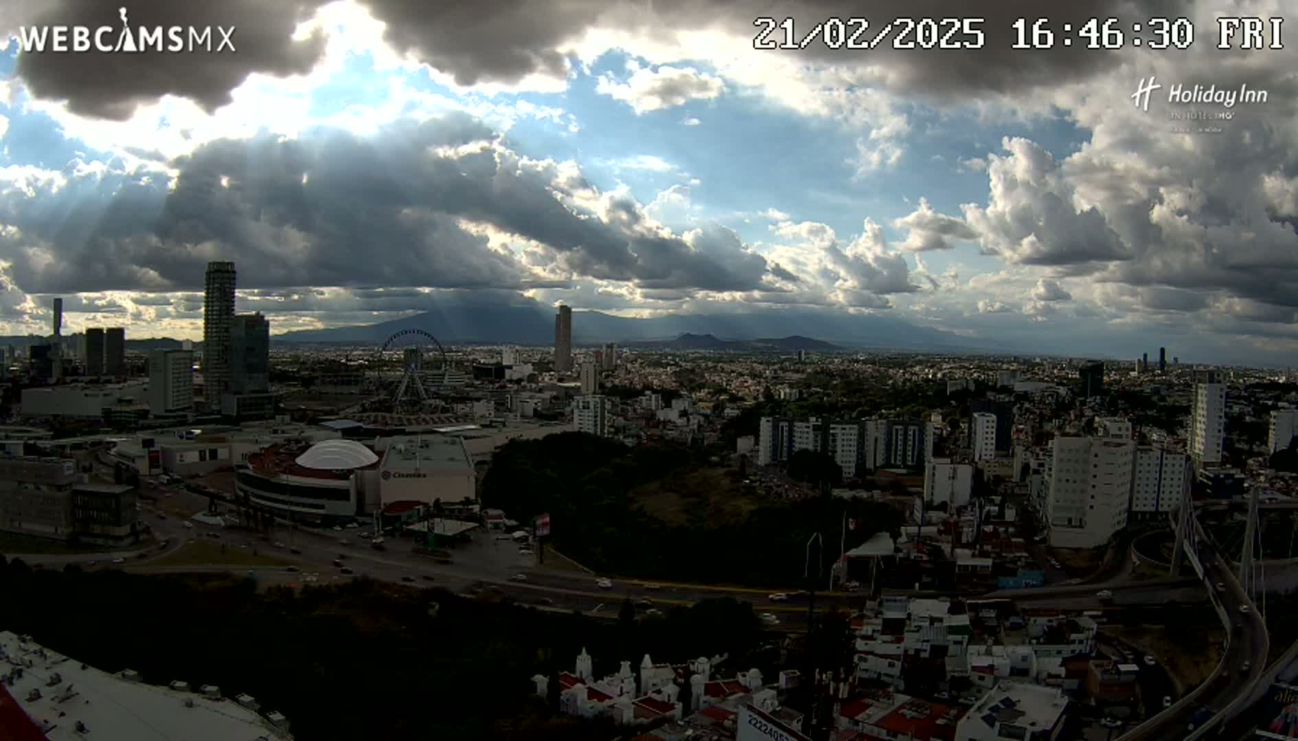 A partly cloudy sky with sunbeams shining through the clouds illuminates a sprawling city with numerous buildings, a Ferris wheel, and distant mountains.