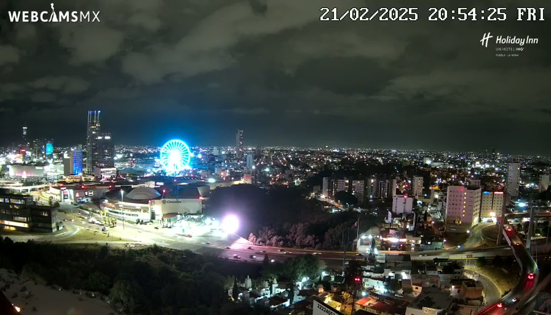 A nighttime, partly cloudy view of a city with a brightly lit Ferris wheel, a large shopping mall, and several high-rise buildings.