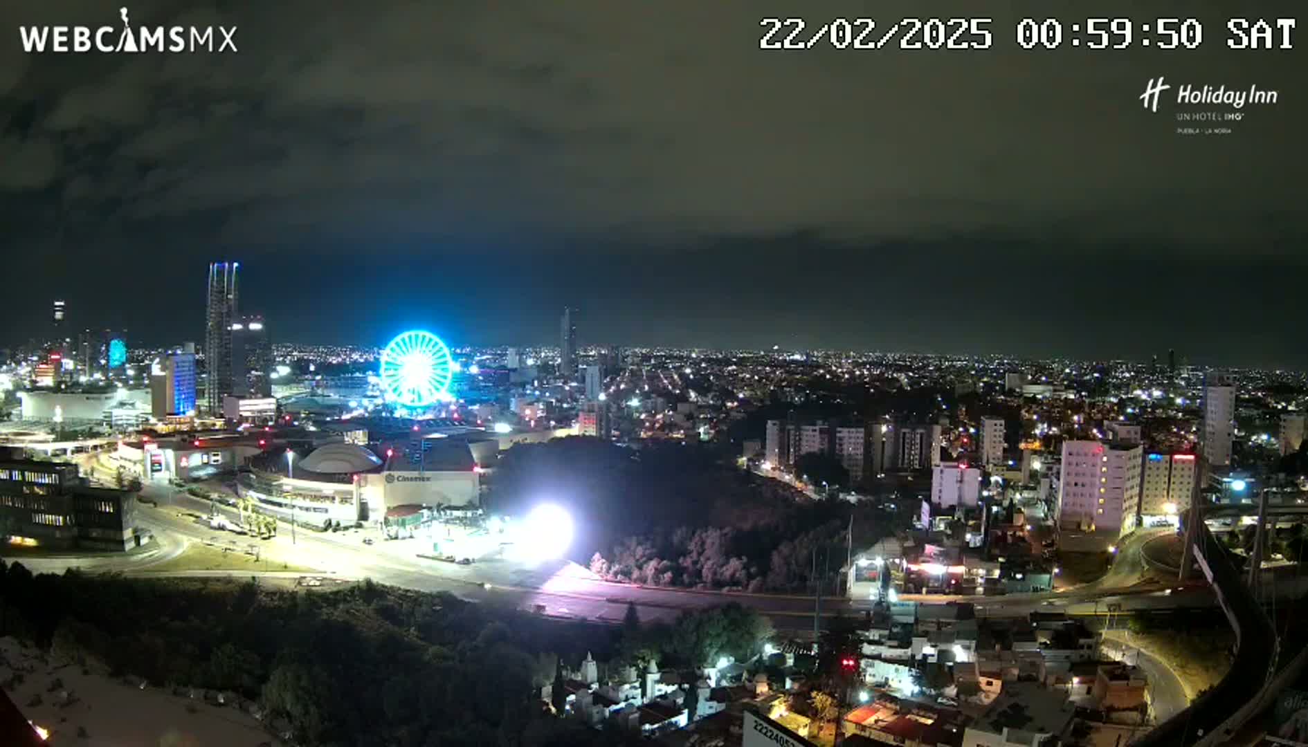 A nighttime cityscape with a Ferris wheel, illuminated buildings, and partly cloudy skies is seen from an elevated vantage point.