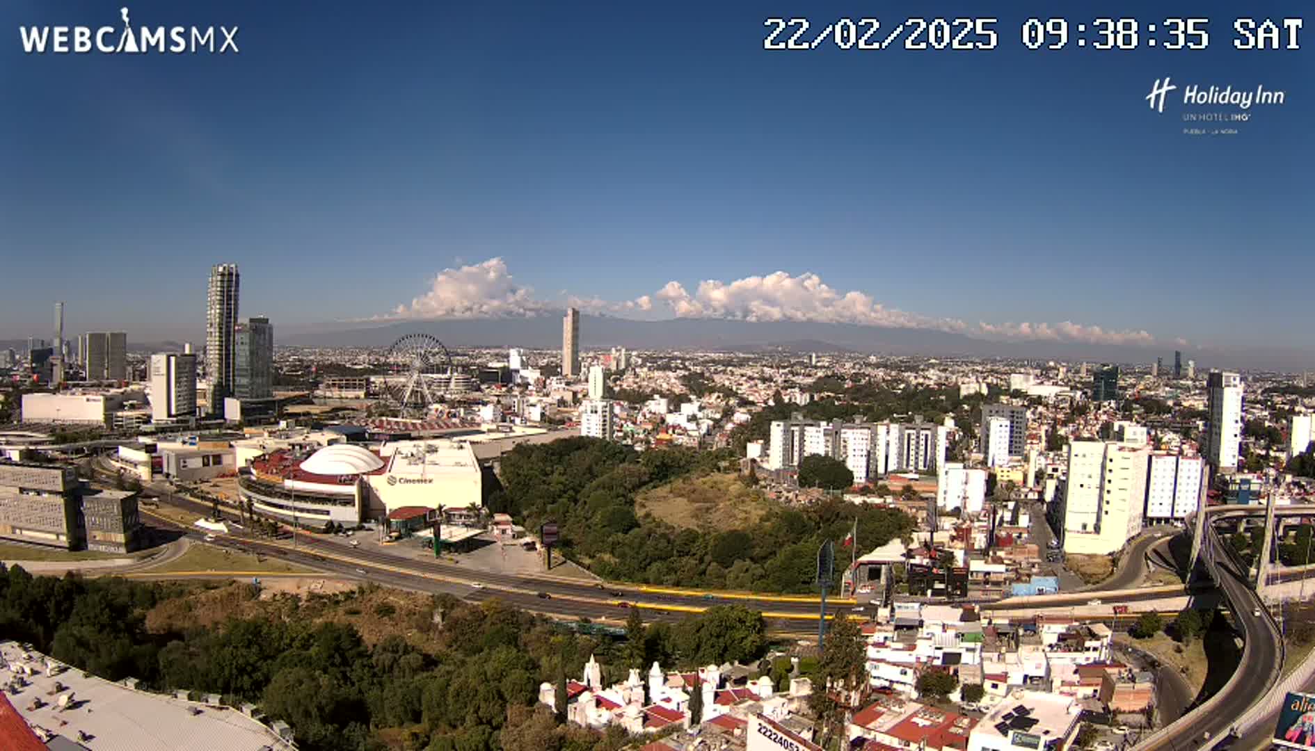 An aerial view of a city on a partly cloudy day, showing a large shopping mall, Ferris wheel, and high-rise buildings, with mountains in the distance.