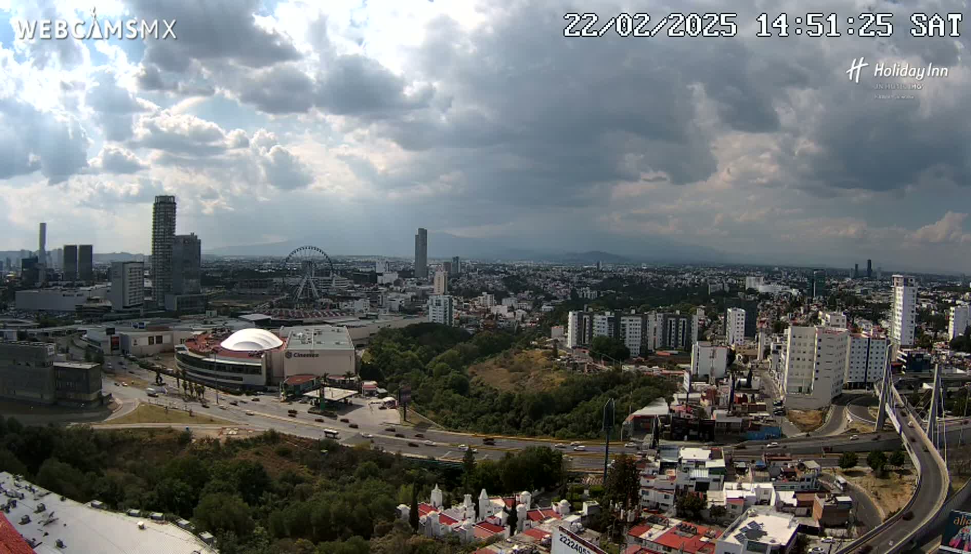 A partly cloudy day reveals a sprawling cityscape with high-rise buildings, a Ferris wheel, and a large shopping mall, all connected by roads and bridges.