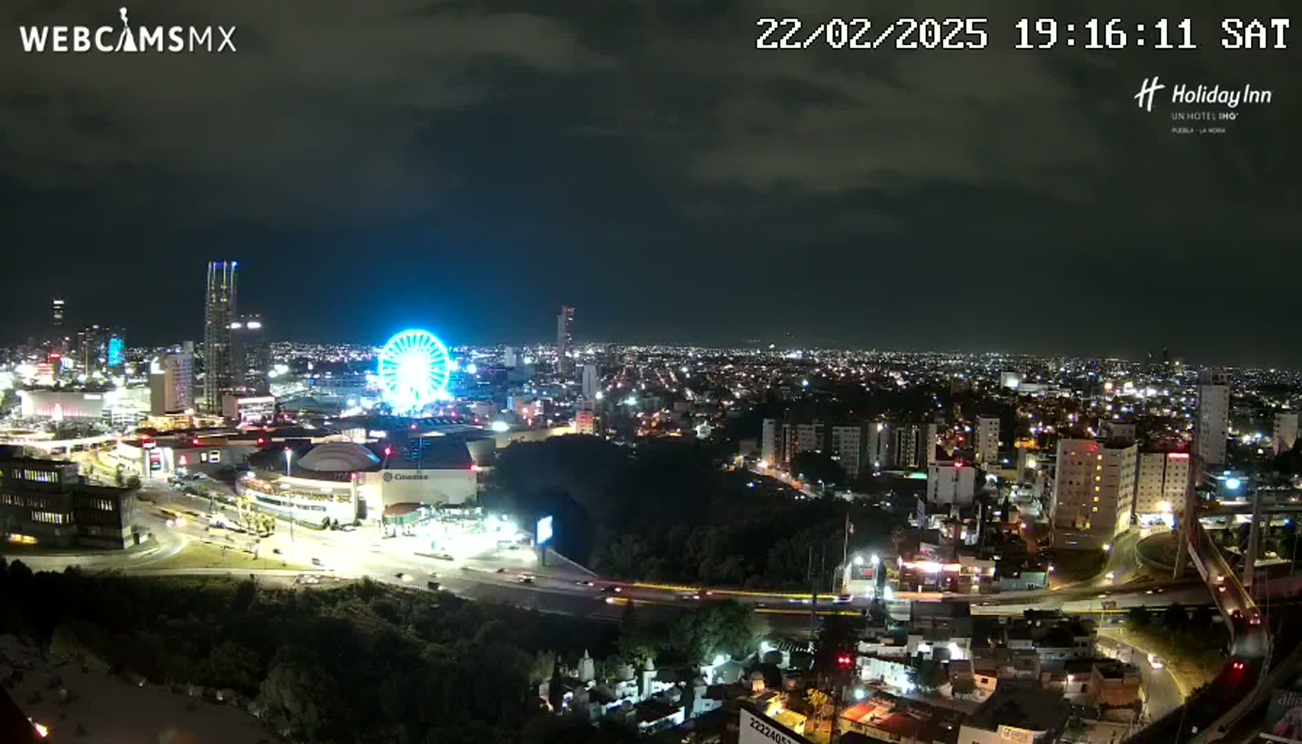 A nighttime view of a city illuminated with lights, featuring a Ferris wheel and a large building complex, under a partly cloudy sky.
