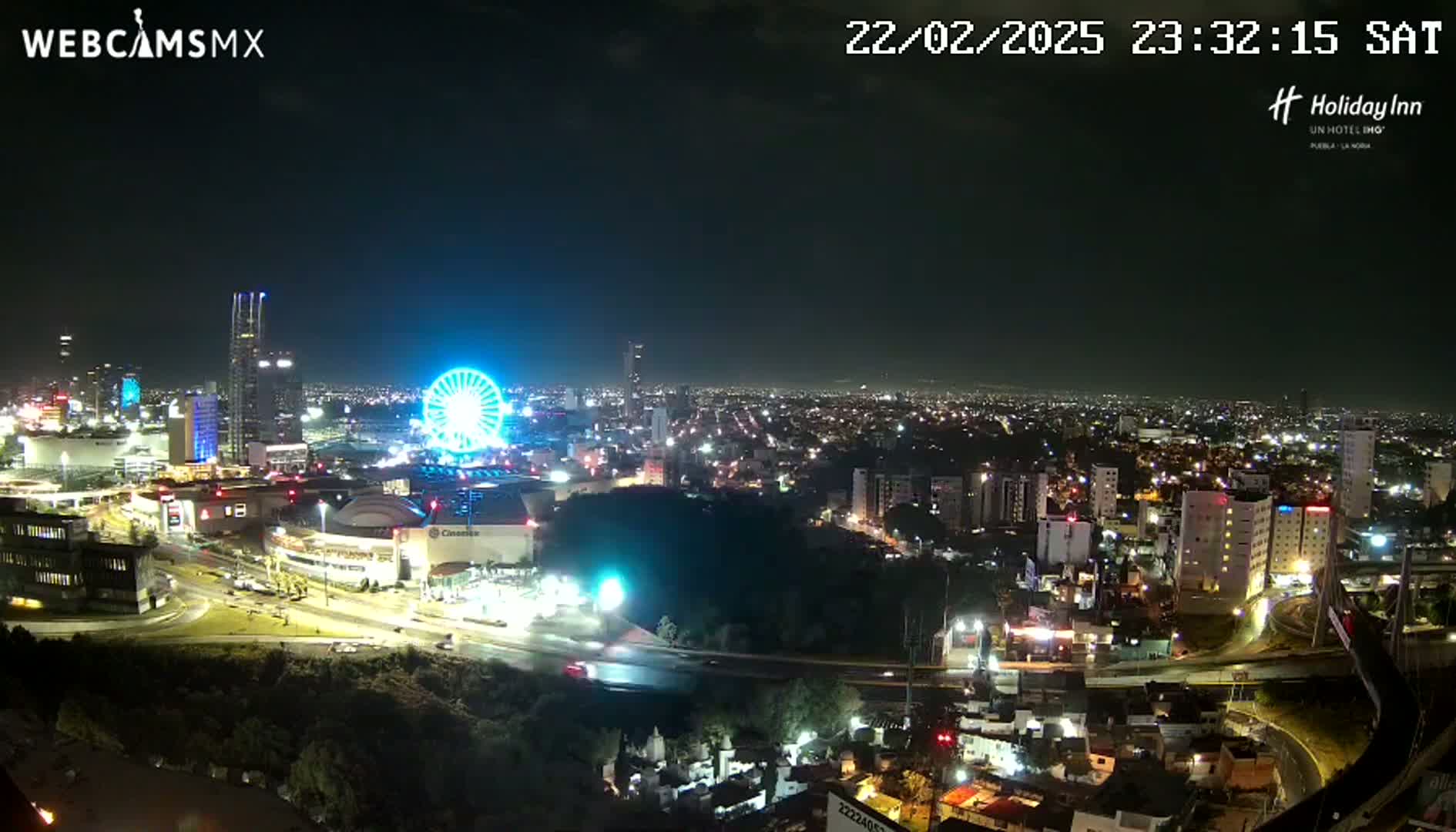 A nighttime, aerial view of a city illuminated with lights, featuring a Ferris wheel and several buildings, under a clear sky.