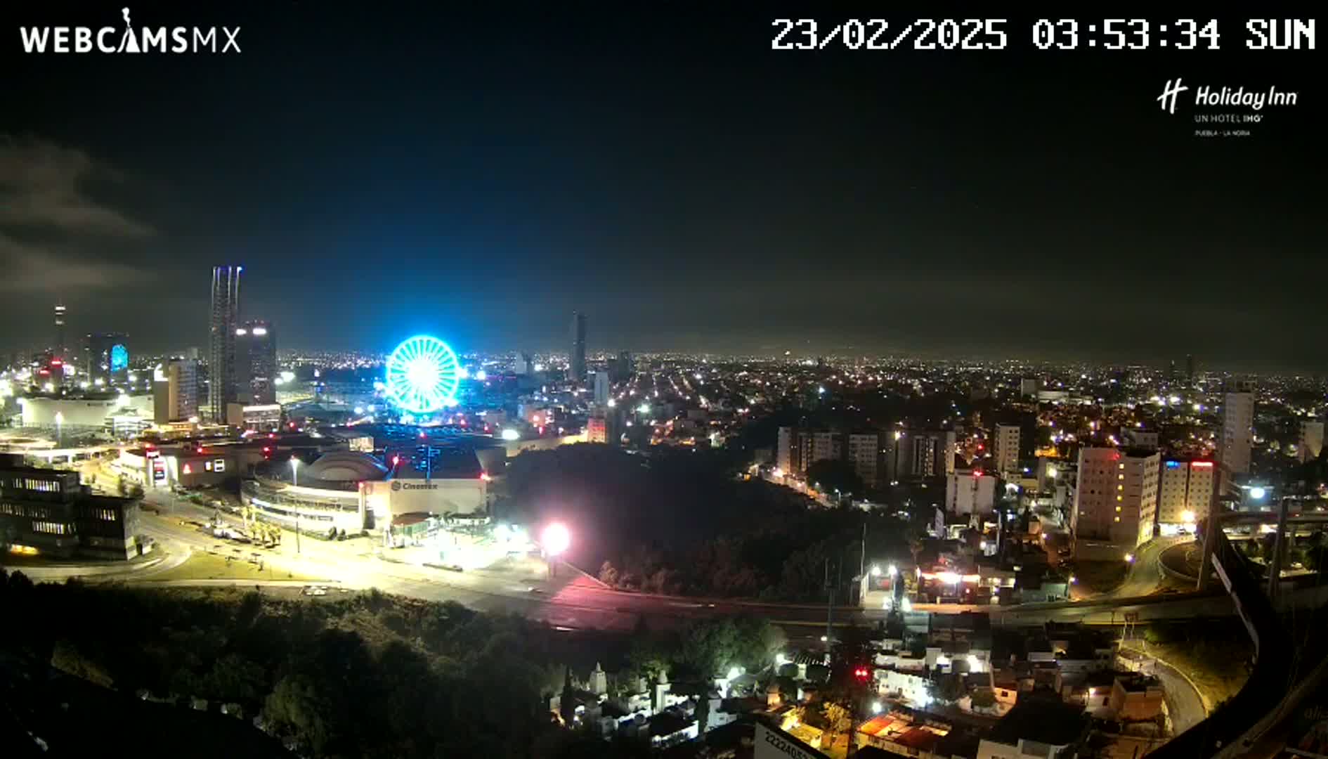A nighttime aerial view of a city illuminated by lights, featuring a Ferris wheel and various buildings, under a mostly clear sky.