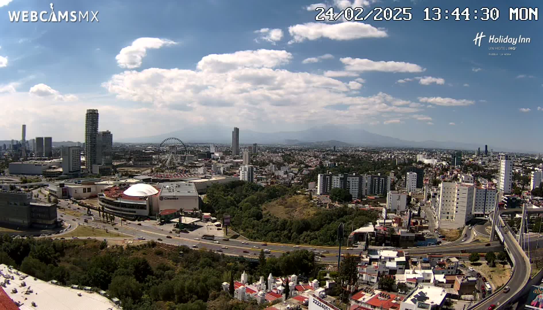 A sprawling city scene under a partly cloudy sky shows a Ferris wheel, several high-rise buildings, and a large shopping mall, all surrounded by dense residential areas and winding roads.