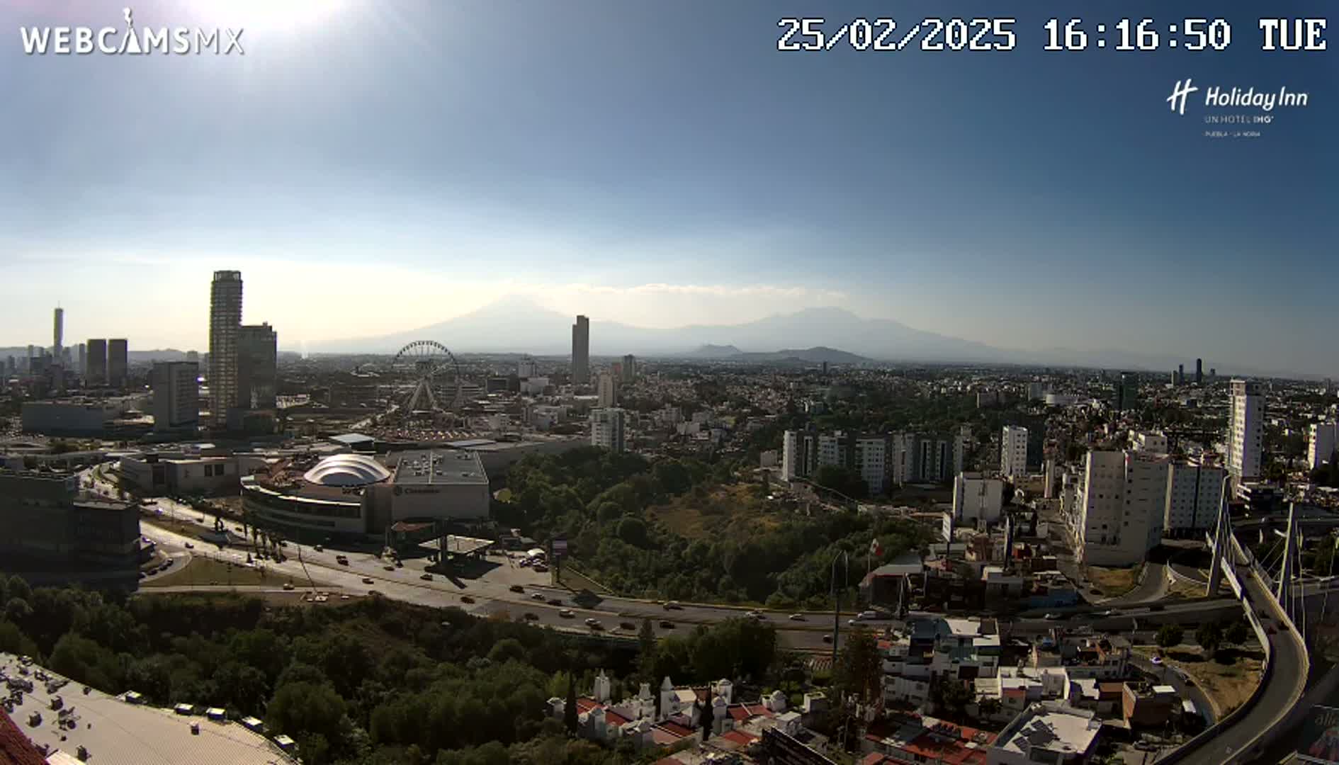 A sunny, high-angle view of a sprawling city with a Ferris wheel, numerous buildings, and mountains in the distance.