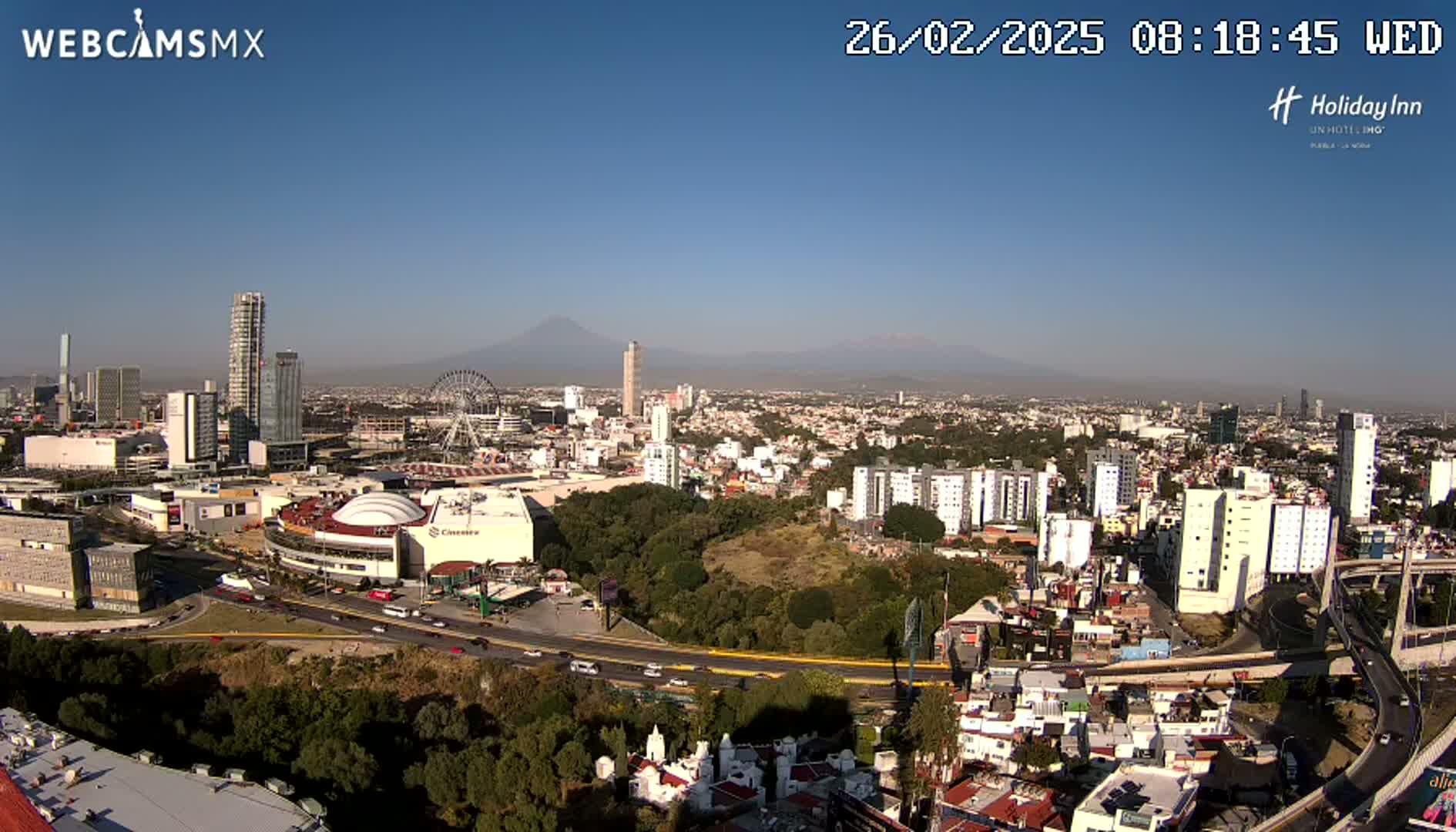 A sprawling cityscape with a Ferris wheel and a distant volcano under clear blue skies.