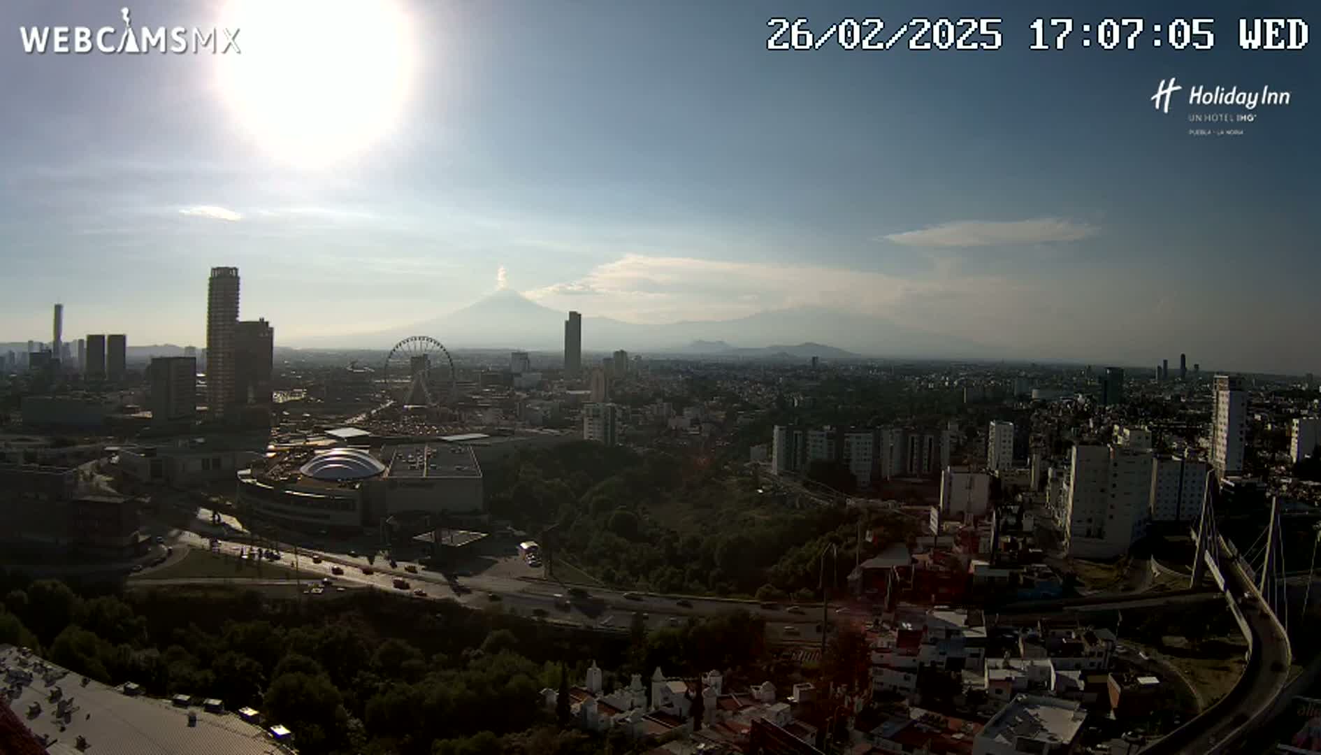 A sunny cityscape with a Ferris wheel and a distant volcano is seen from a high vantage point.