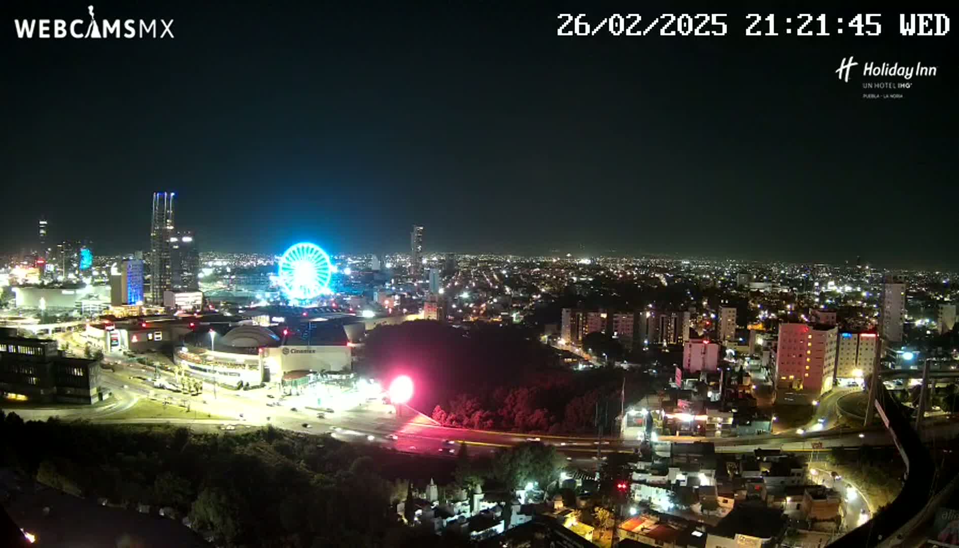 A nighttime view of a city illuminated with lights, featuring a Ferris wheel and a large shopping mall, under a clear sky.