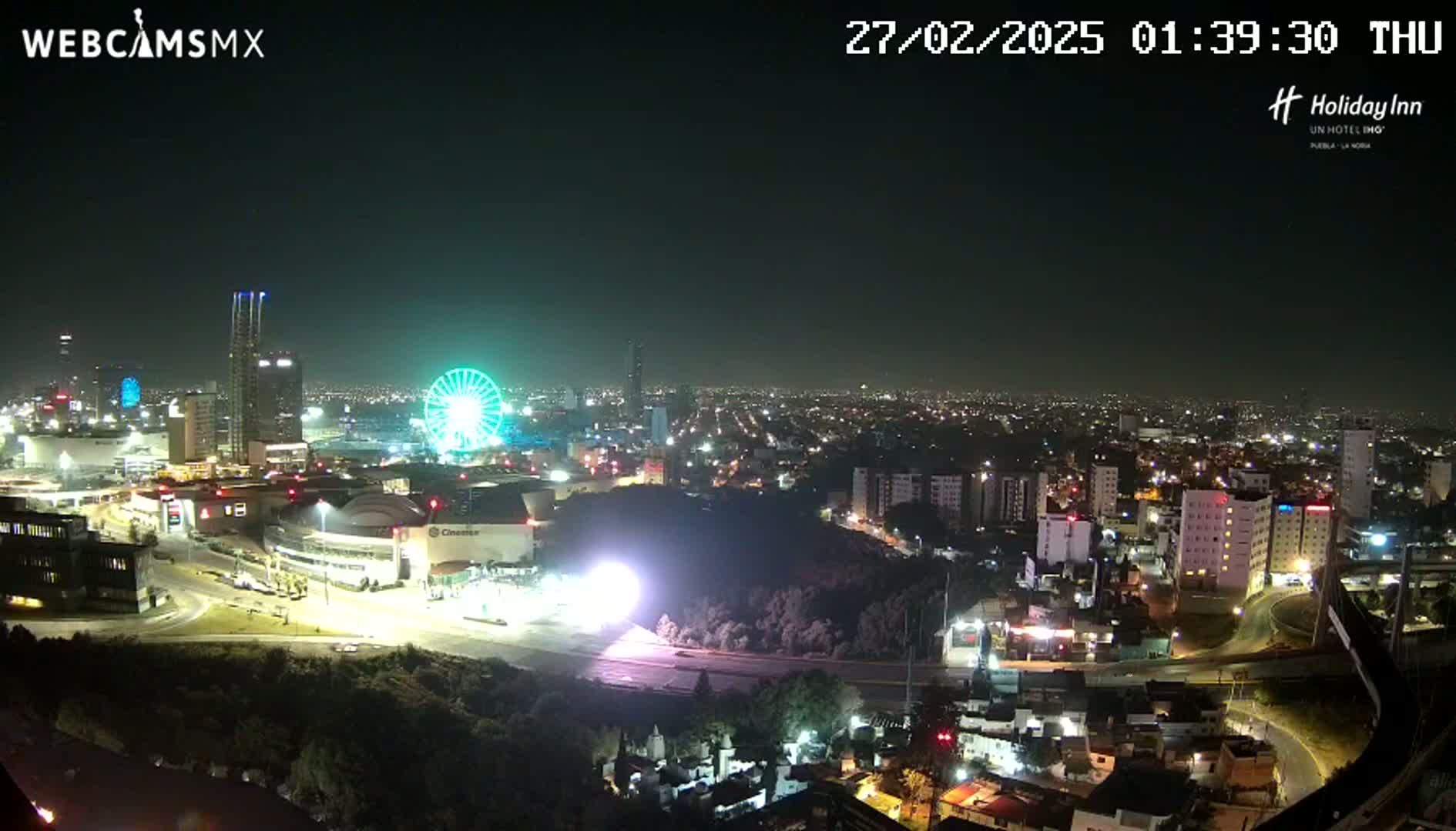 A nighttime, aerial view of a city illuminated by lights, featuring a Ferris wheel and several high-rise buildings.