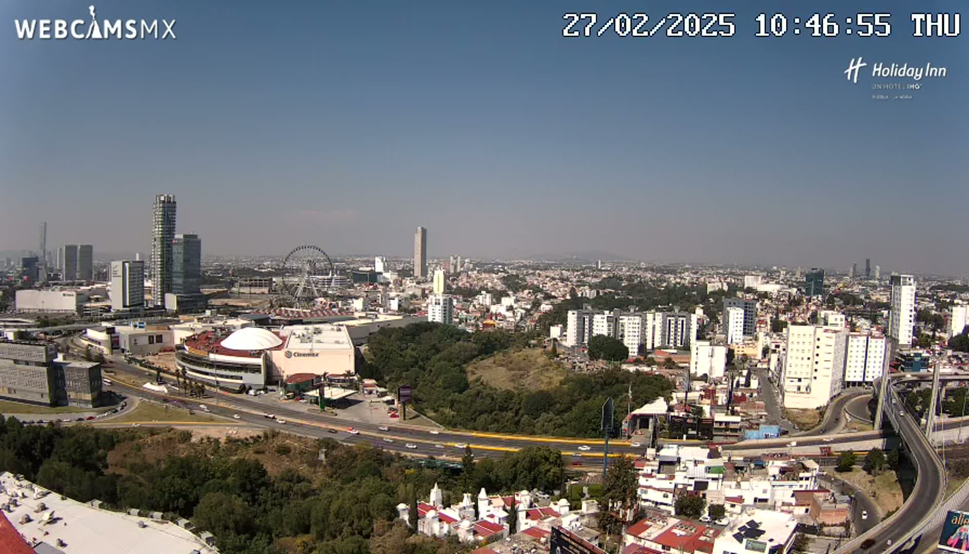 A panoramic view of a city on a clear, sunny day, showing a large shopping mall, a Ferris wheel, numerous high-rise buildings, and a network of roads and highways.