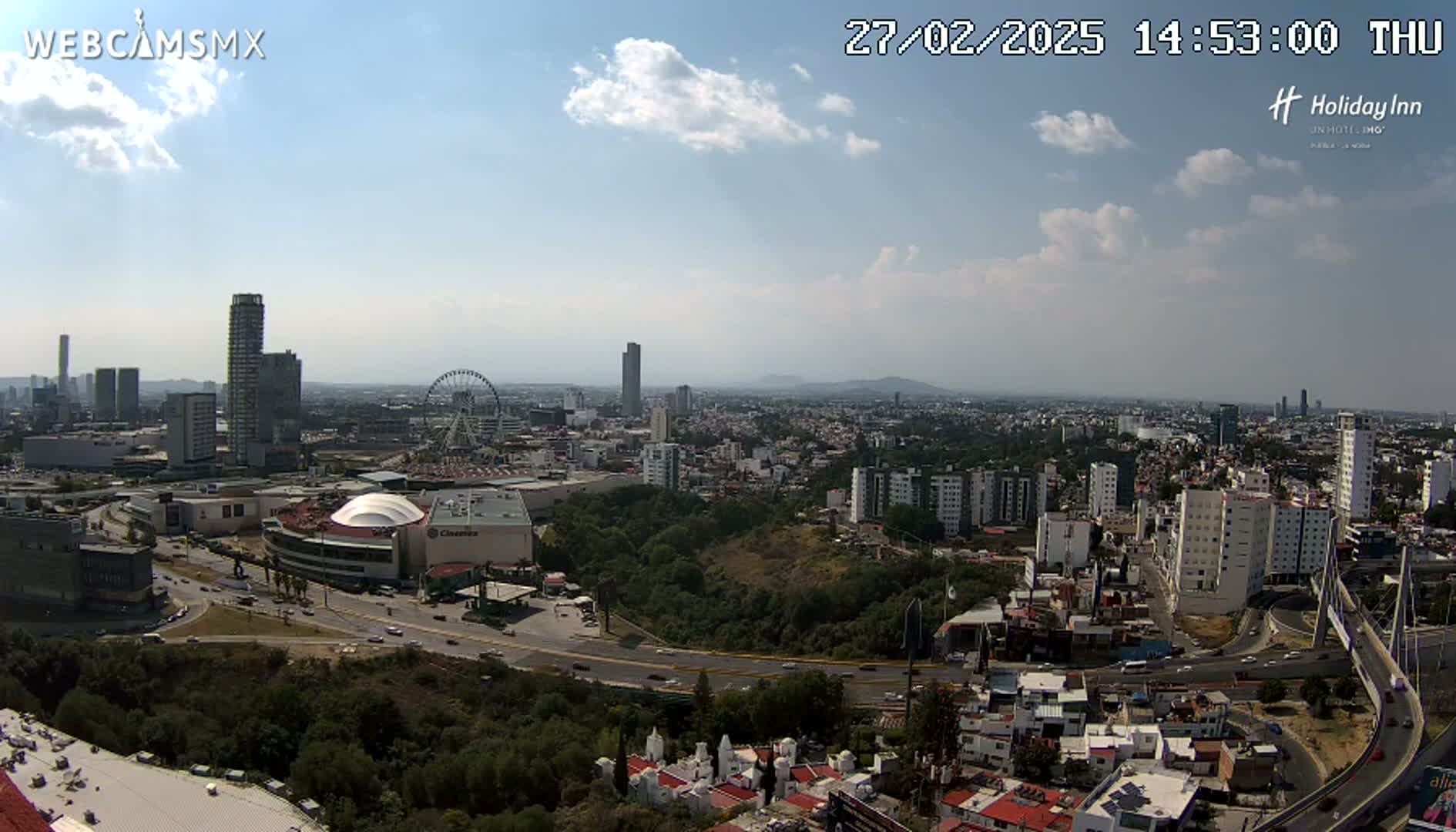 A partly cloudy daytime view of a sprawling city with tall buildings, a Ferris wheel, and a large dome-shaped structure, seen from an elevated vantage point overlooking a highway and surrounding hills.