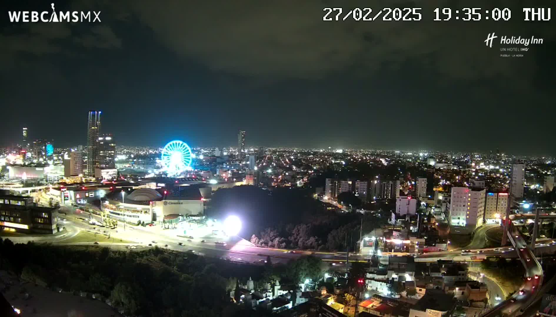 A nighttime view of a city with a Ferris wheel and illuminated buildings under a partly cloudy sky.