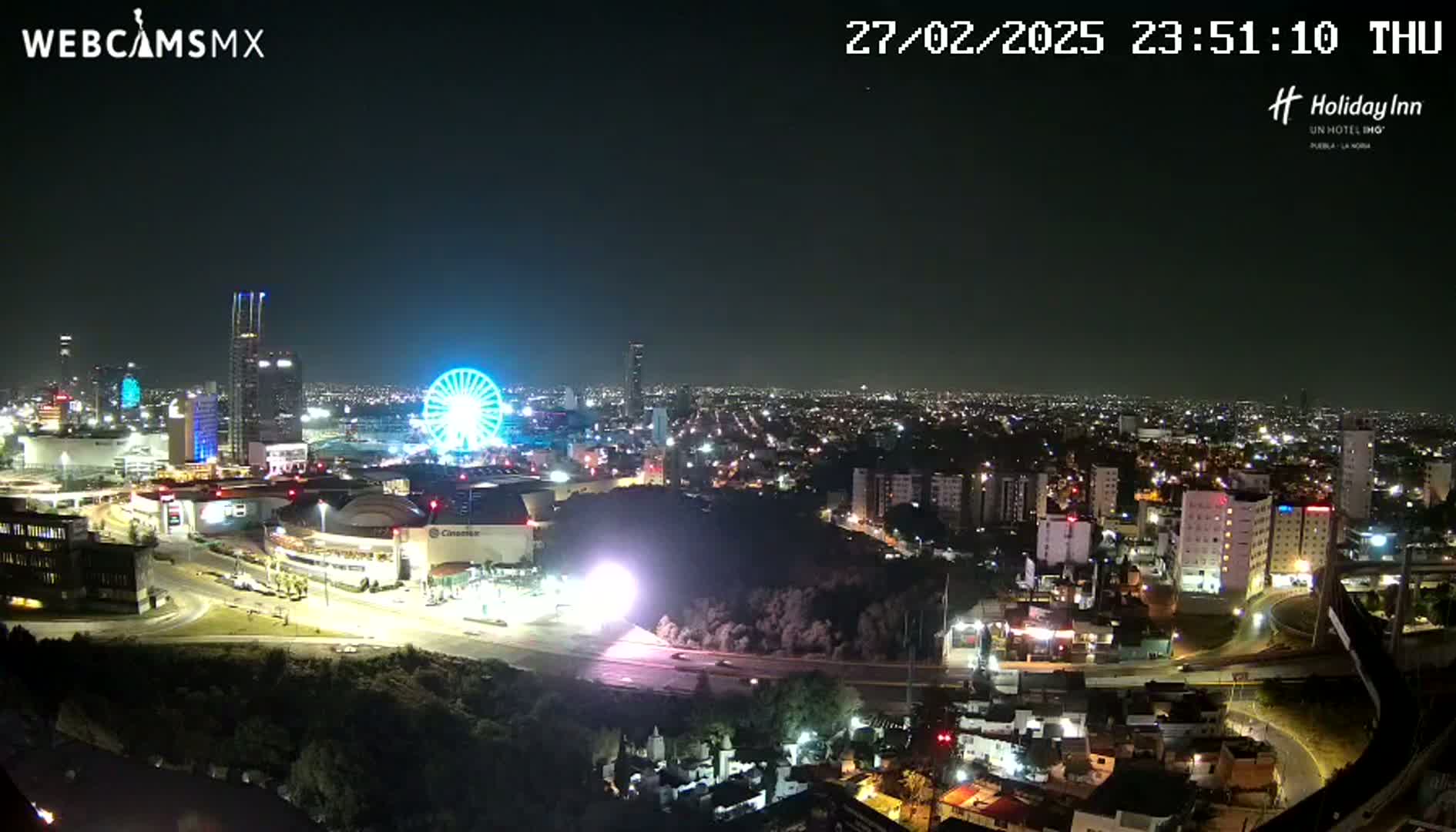 A nighttime aerial view of a city illuminated by lights, featuring a Ferris wheel and a large shopping center complex, under a clear sky.