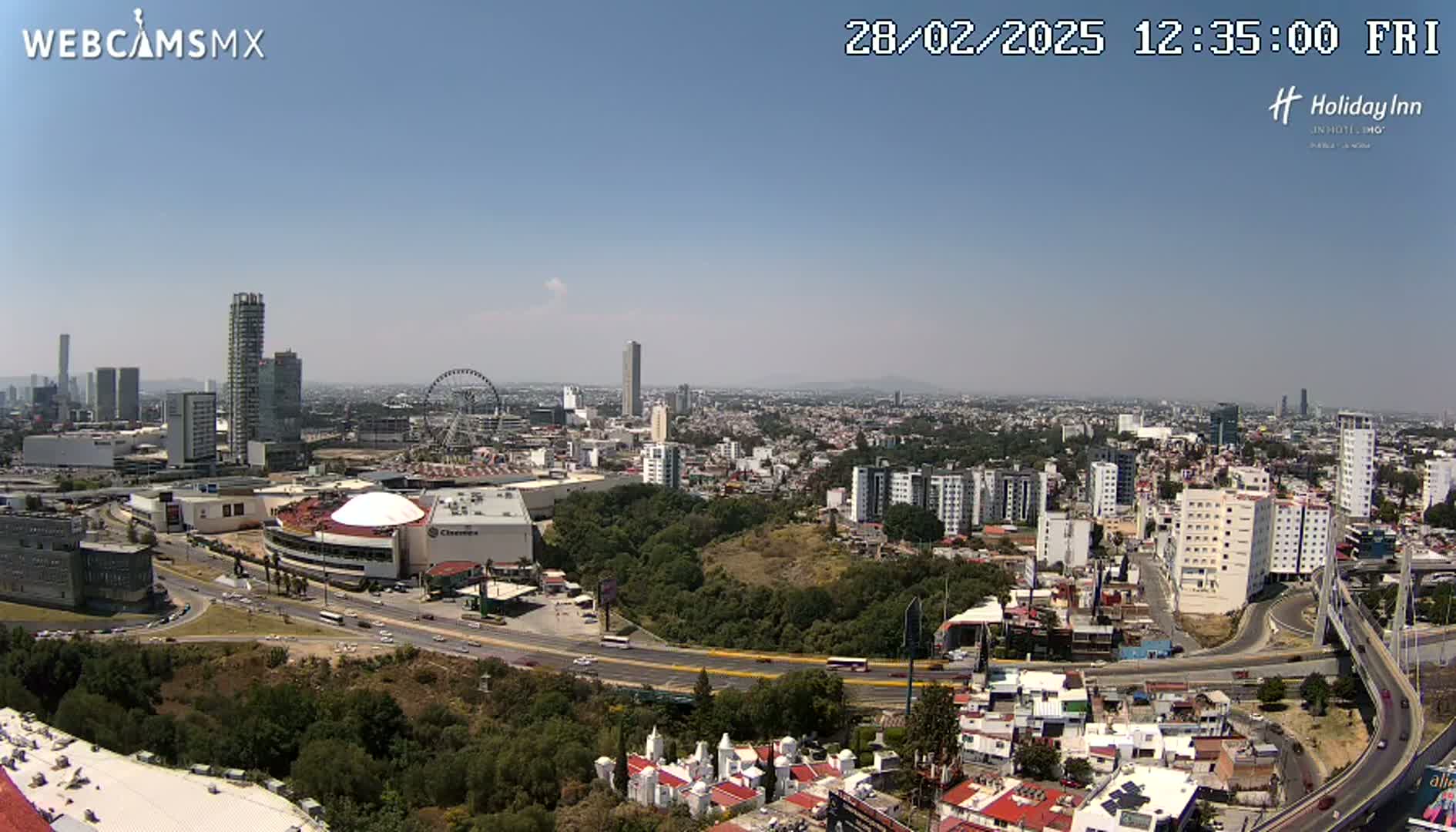 A sprawling city scene under a clear, sunny sky shows a Ferris wheel, several high-rise buildings, and a large shopping center next to a highway.