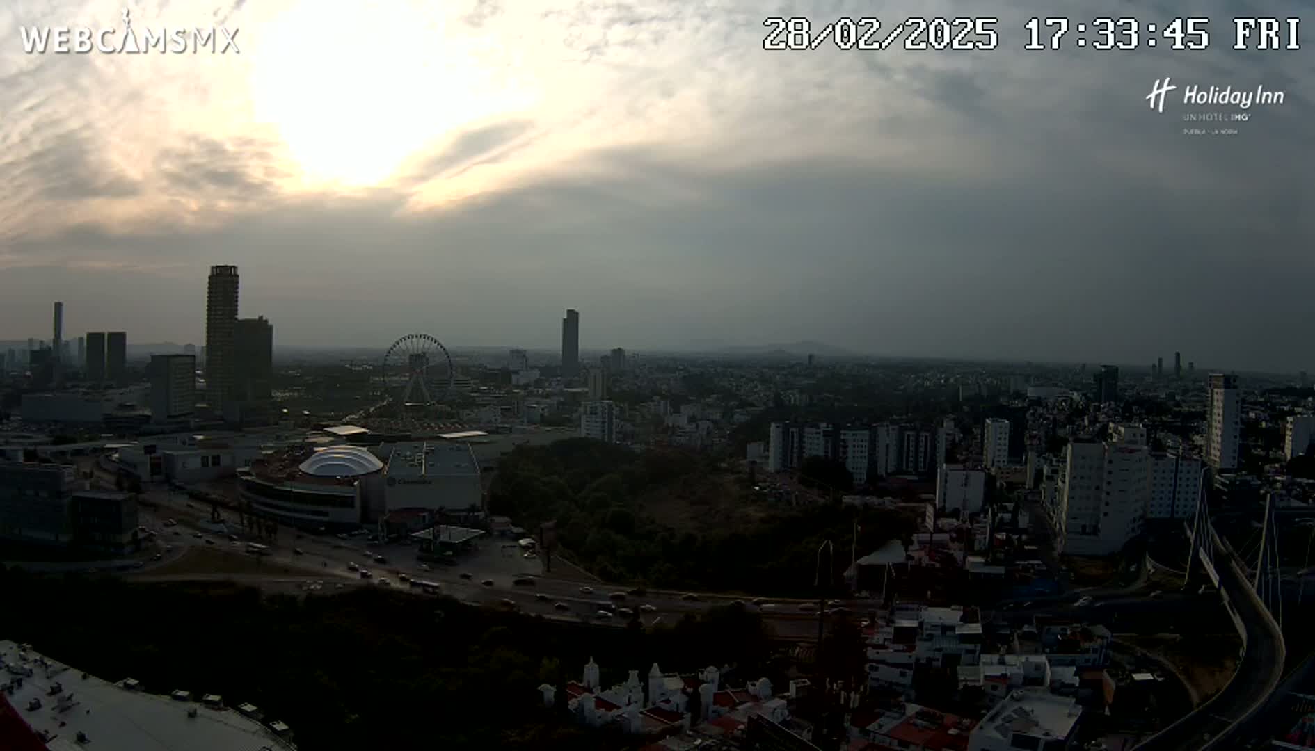 A hazy sunset casts a dim light over a sprawling city, showcasing a Ferris wheel and numerous buildings, with a partially cloudy sky overhead.