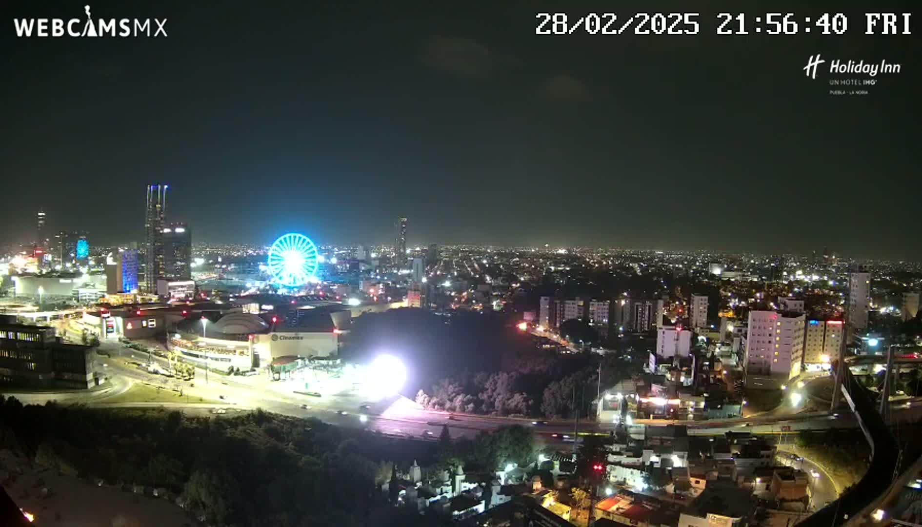 A nighttime, city view shows a brightly lit Ferris wheel and a sprawling cityscape under a clear sky.