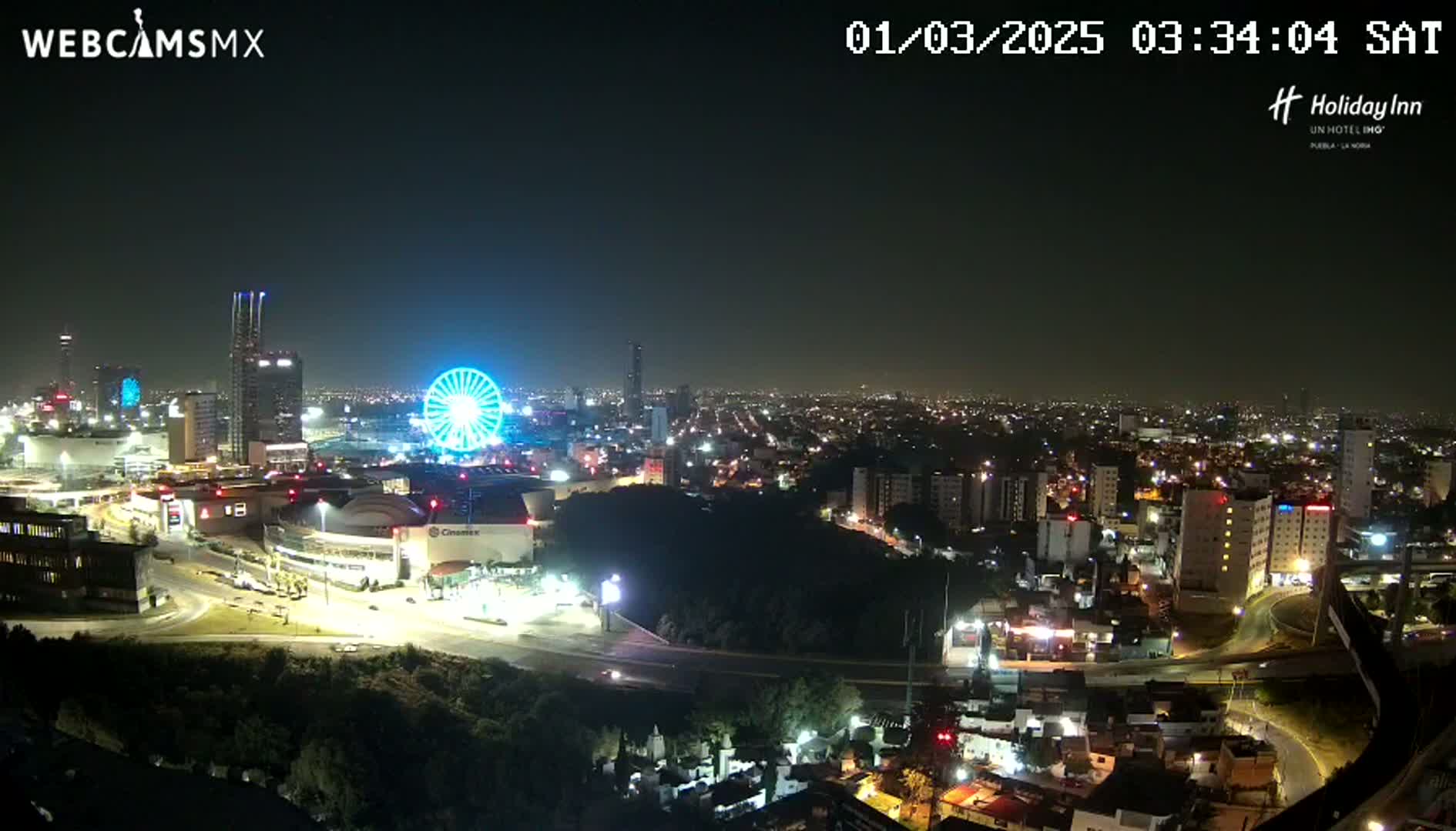 A nighttime, aerial view of a city with a brightly lit Ferris wheel and numerous buildings, under a clear sky.
