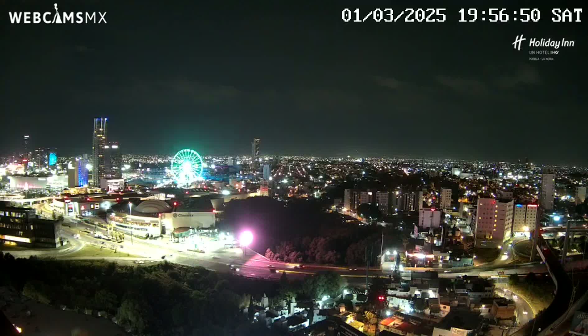 A nighttime, aerial view of a city with a Ferris wheel and several buildings illuminated, under a clear sky.