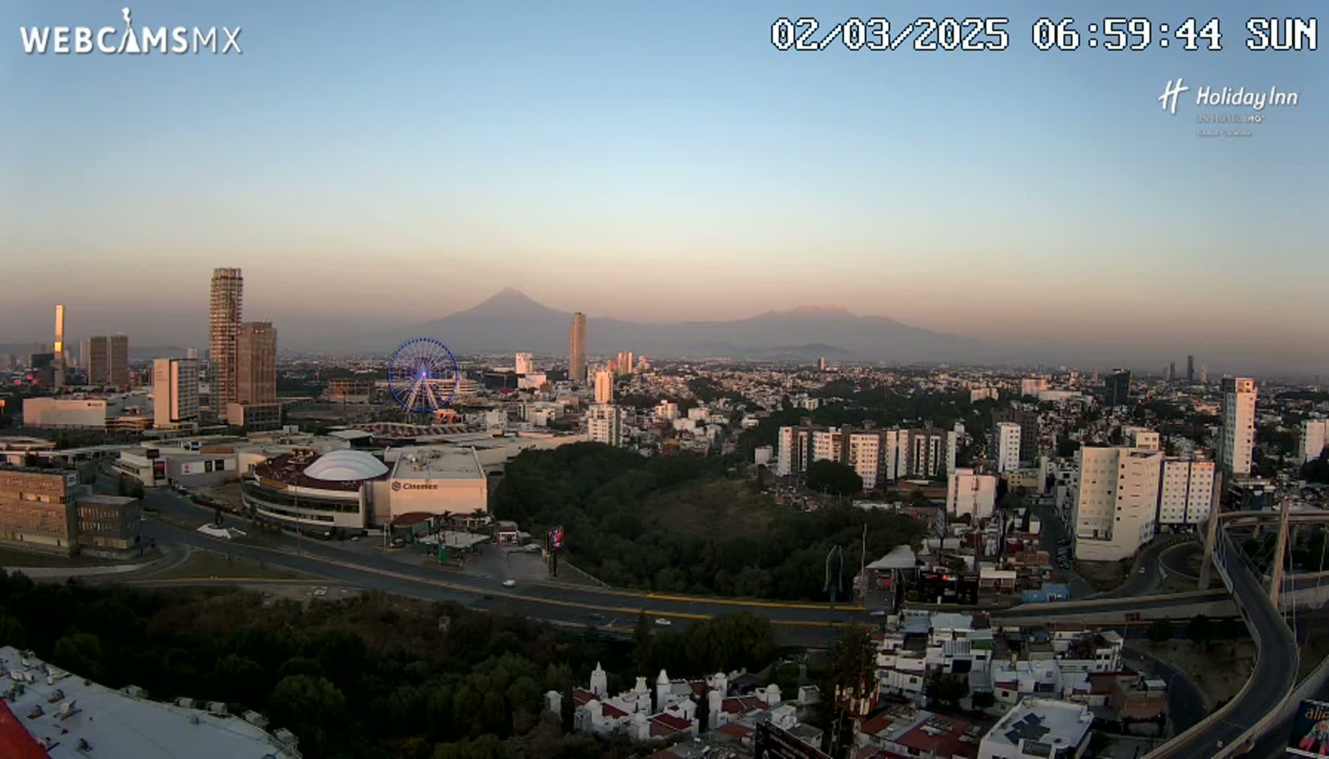 A panoramic view of a city at sunrise, featuring a Ferris wheel, numerous buildings, and mountains in the distance under a clear sky.