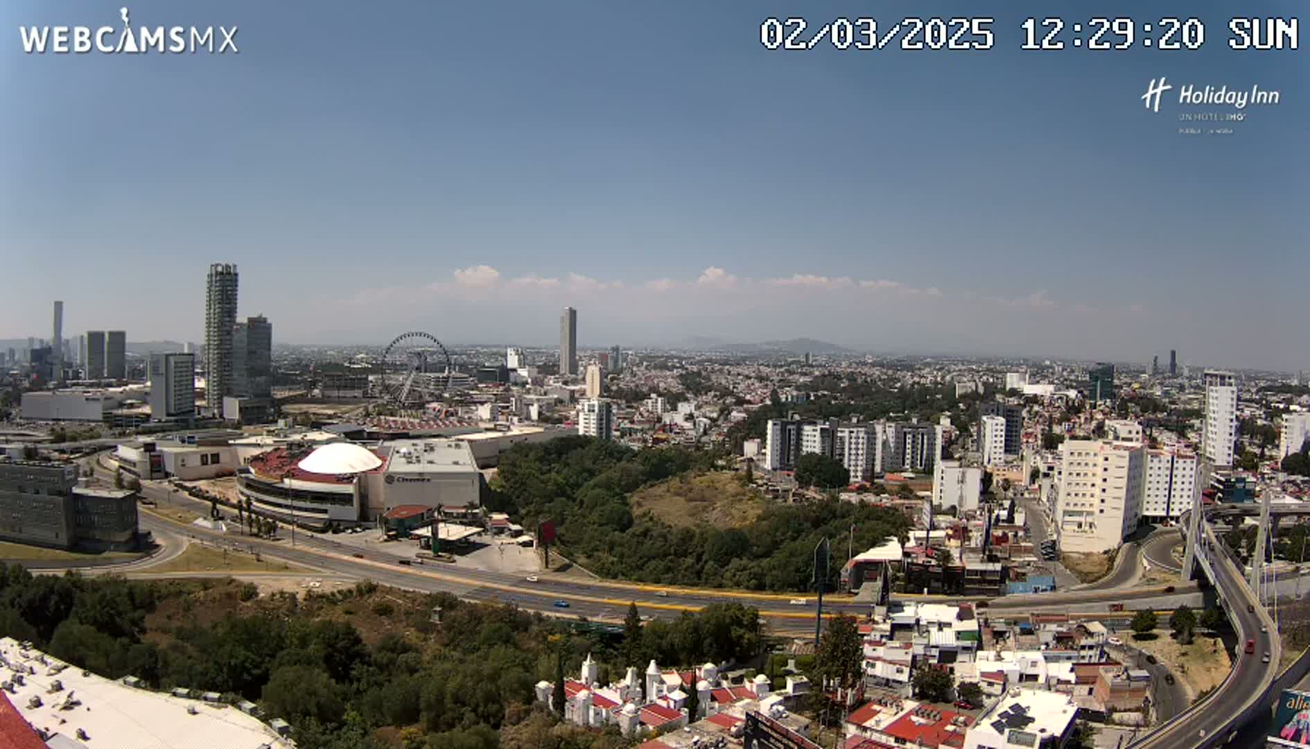 A sprawling cityscape under a mostly clear, sunny sky, featuring high-rise buildings, a large shopping mall with a dome, a Ferris wheel, and winding roads.