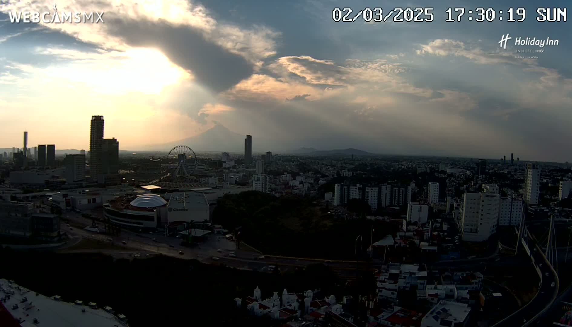 A partly cloudy sunset over a sprawling city with a Ferris wheel and a volcano visible in the distance.