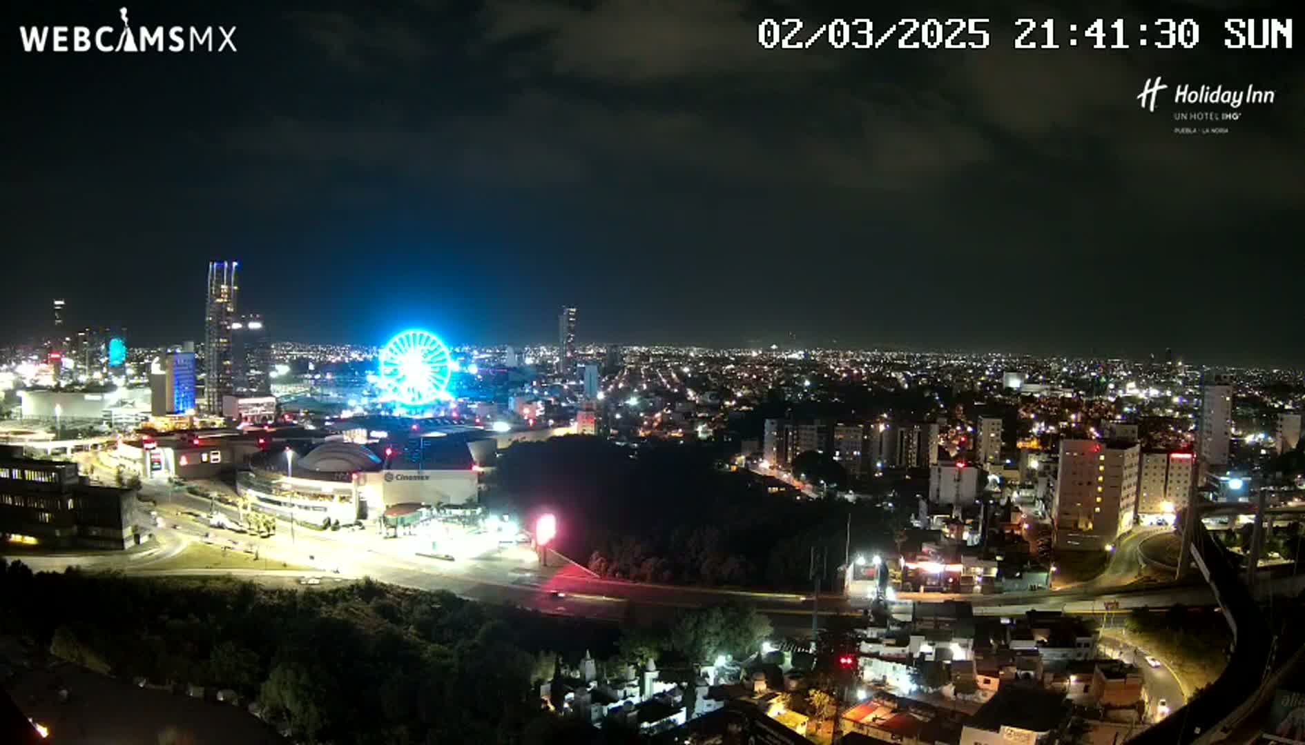 A nighttime aerial view of a city illuminated with lights, featuring a brightly lit Ferris wheel near a large shopping center.