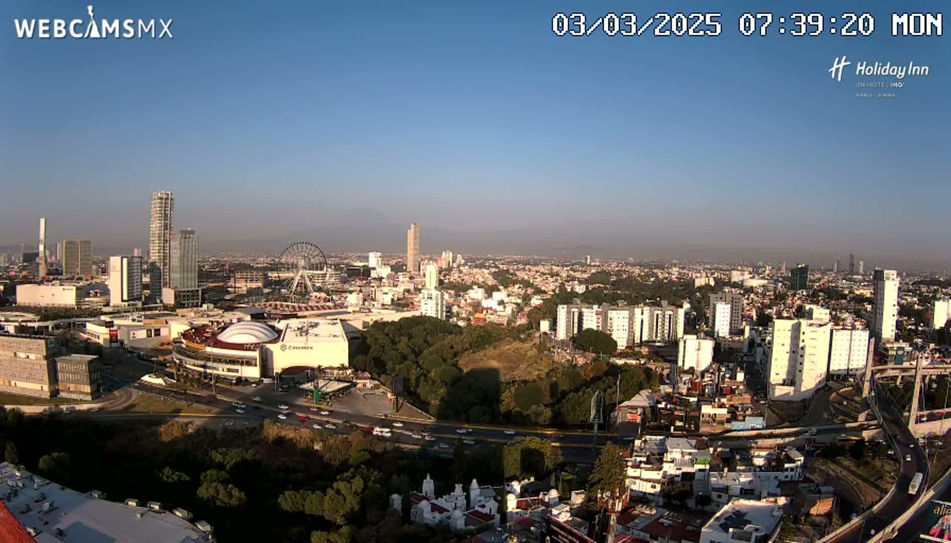 A high-angle, long shot of a city on a clear, sunny day shows a Ferris wheel, a large stadium, and many buildings, with a highway visible in the foreground.
