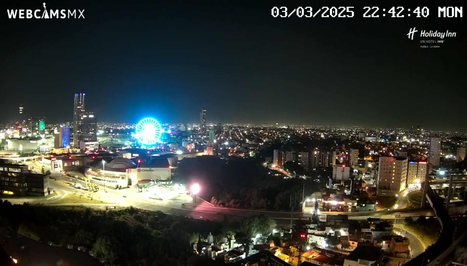 A nighttime, illuminated cityscape view shows a Ferris wheel, shopping center, and numerous buildings extending into the distance under a clear sky.