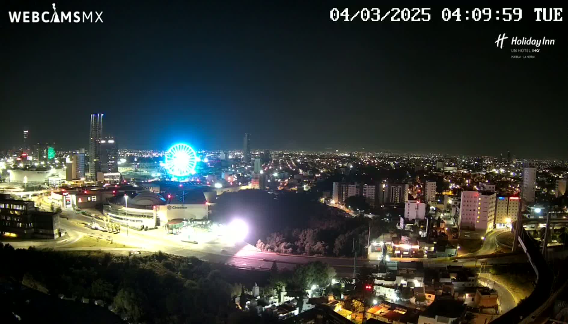 A nighttime aerial view of a city with a brightly lit Ferris wheel near a large building complex, under a clear sky.