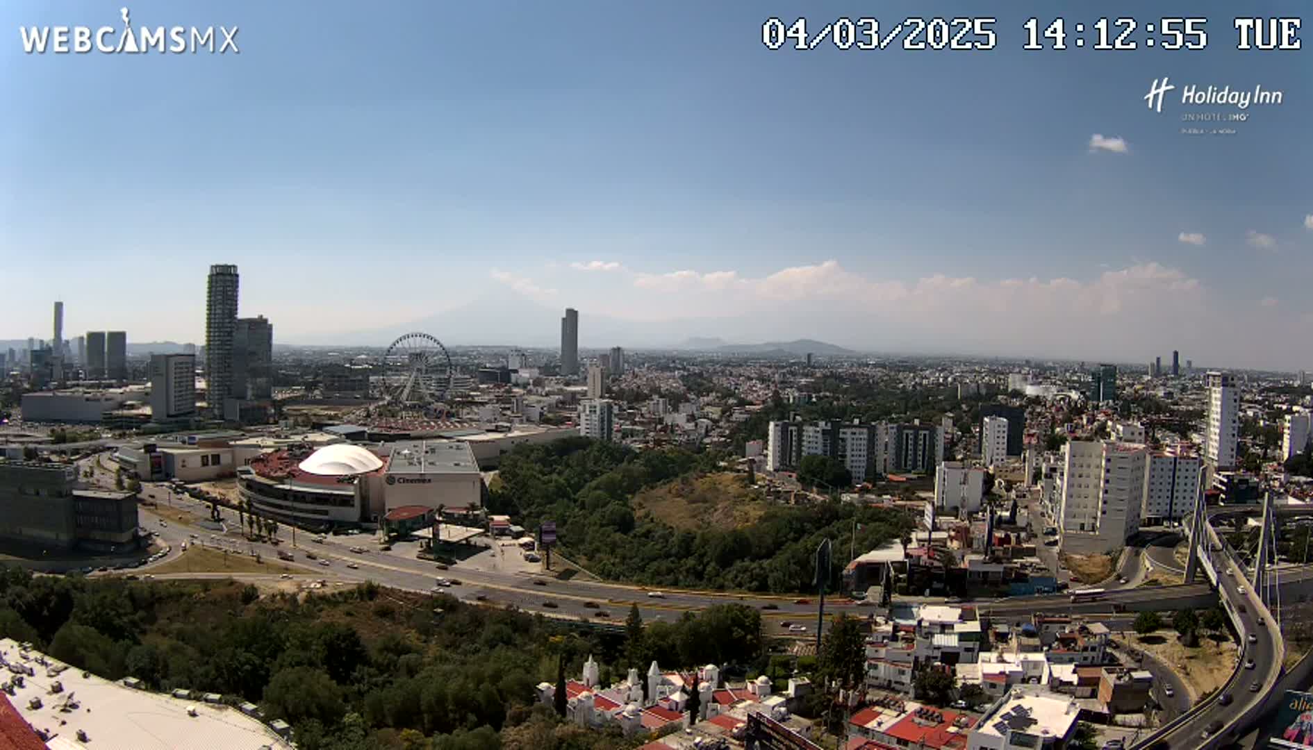 A sprawling city scene under a mostly sunny sky shows a Ferris wheel, numerous buildings, and a highway system with a cable-stayed bridge.