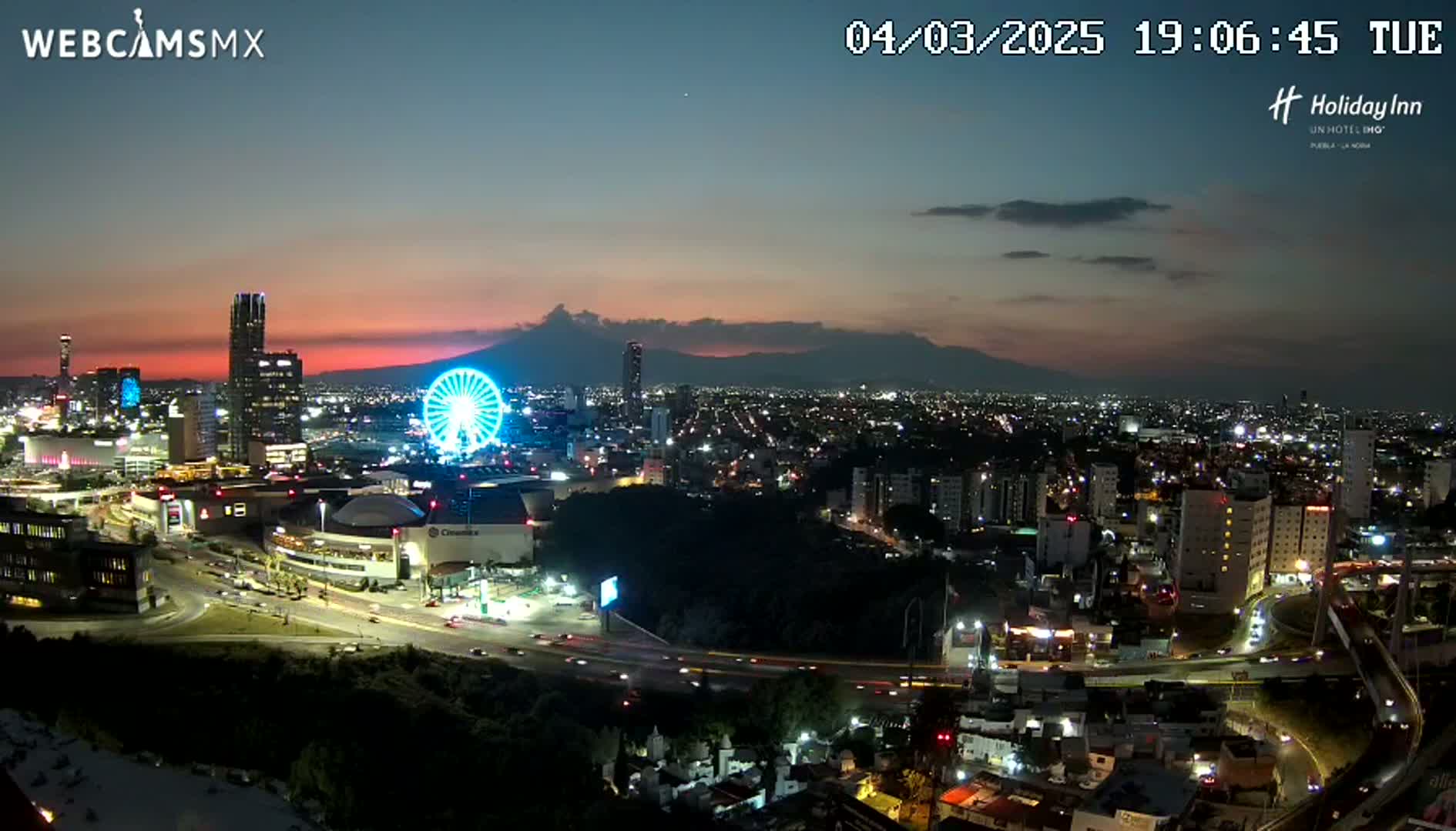 A nighttime, aerial view of a city with a Ferris wheel and illuminated buildings, showcasing a partly cloudy sunset over distant mountains.