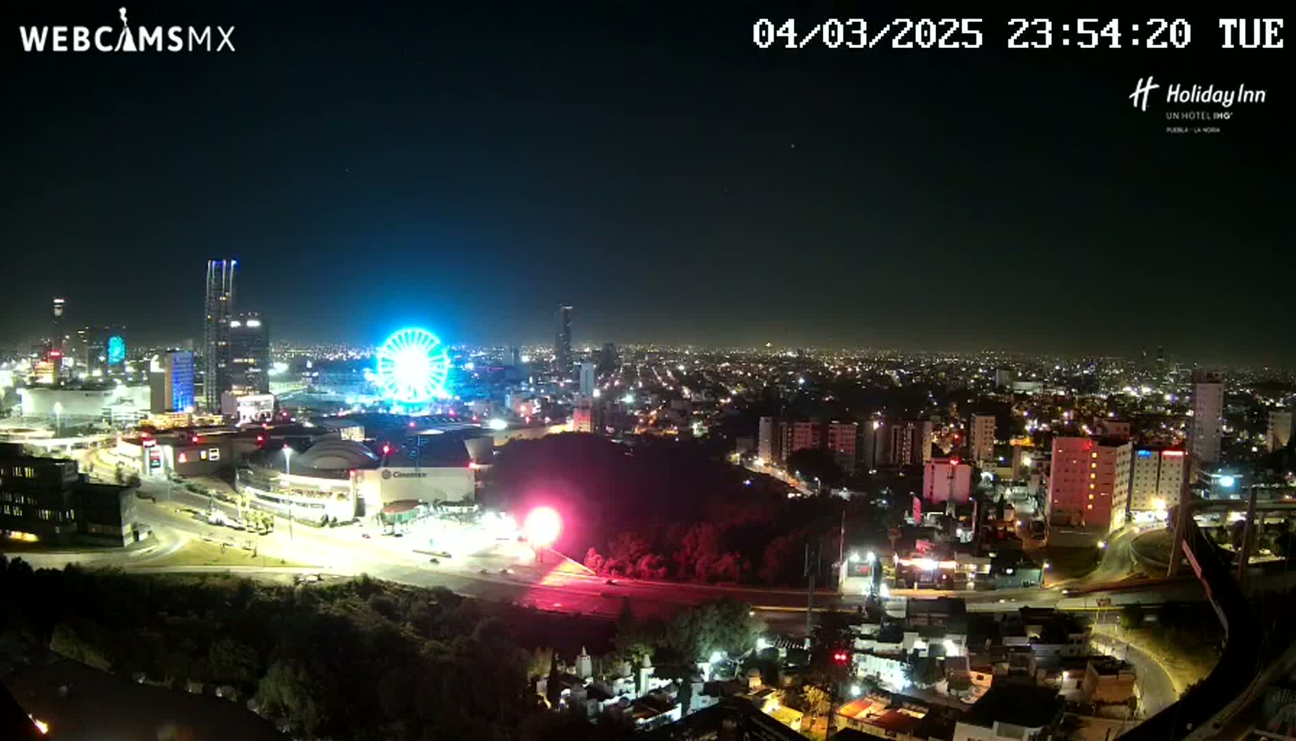 A nighttime, aerial view of a city illuminated by lights, featuring a Ferris wheel and several tall buildings under a clear sky.