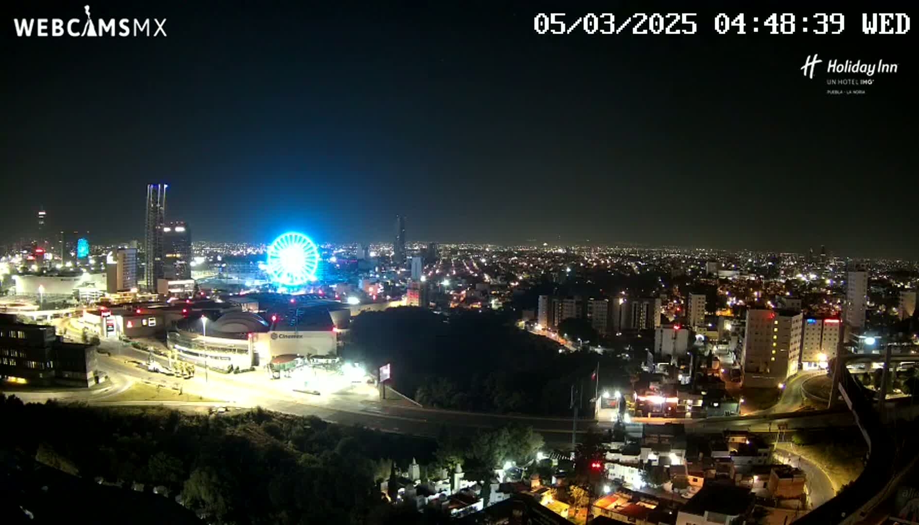 A nighttime aerial view of a city illuminated by lights, featuring a Ferris wheel and a large building complex, under a clear sky.