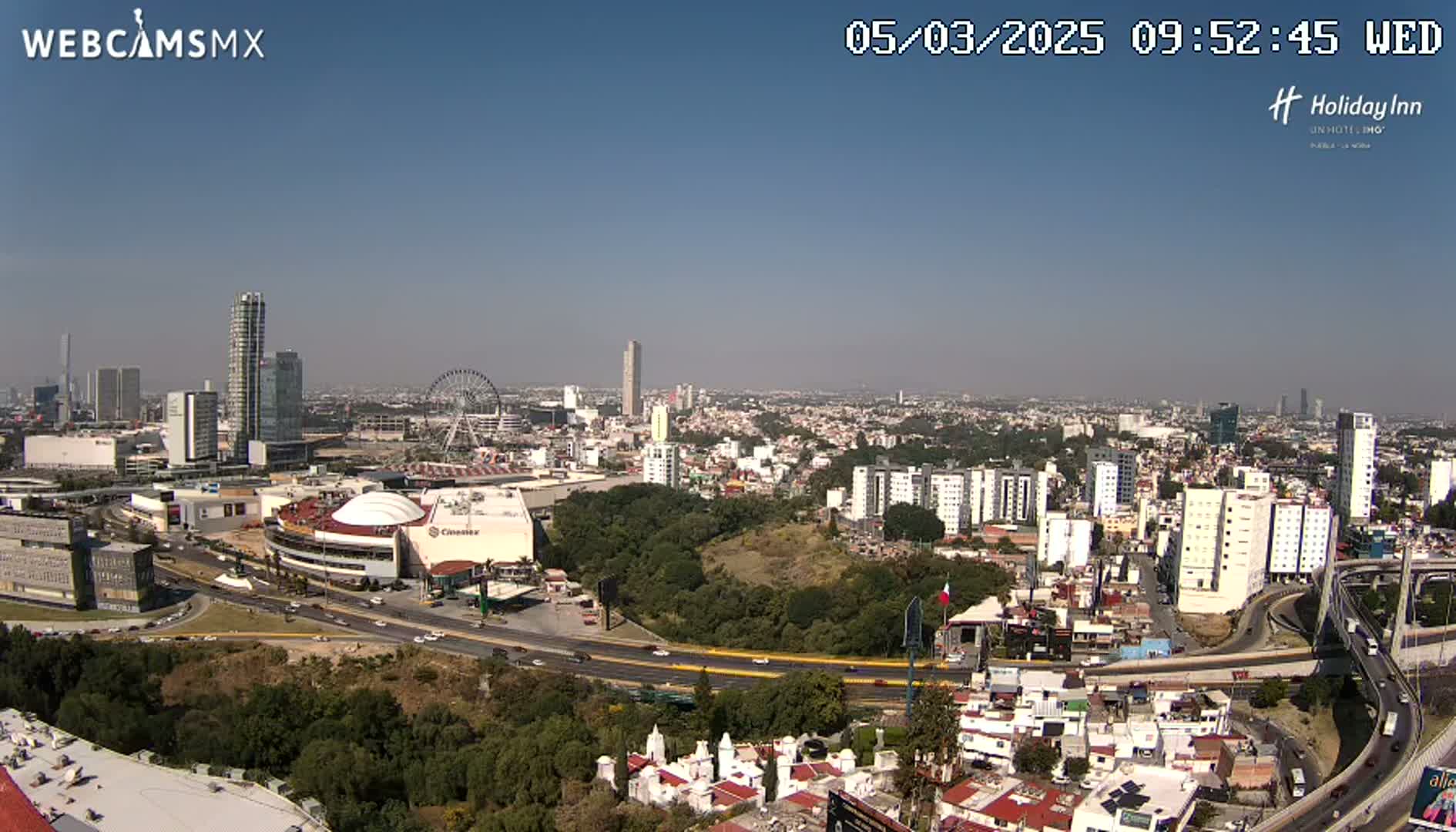 A panoramic view of a city on a clear, sunny day shows a cityscape with numerous buildings, highways, a Ferris wheel, and green spaces.
