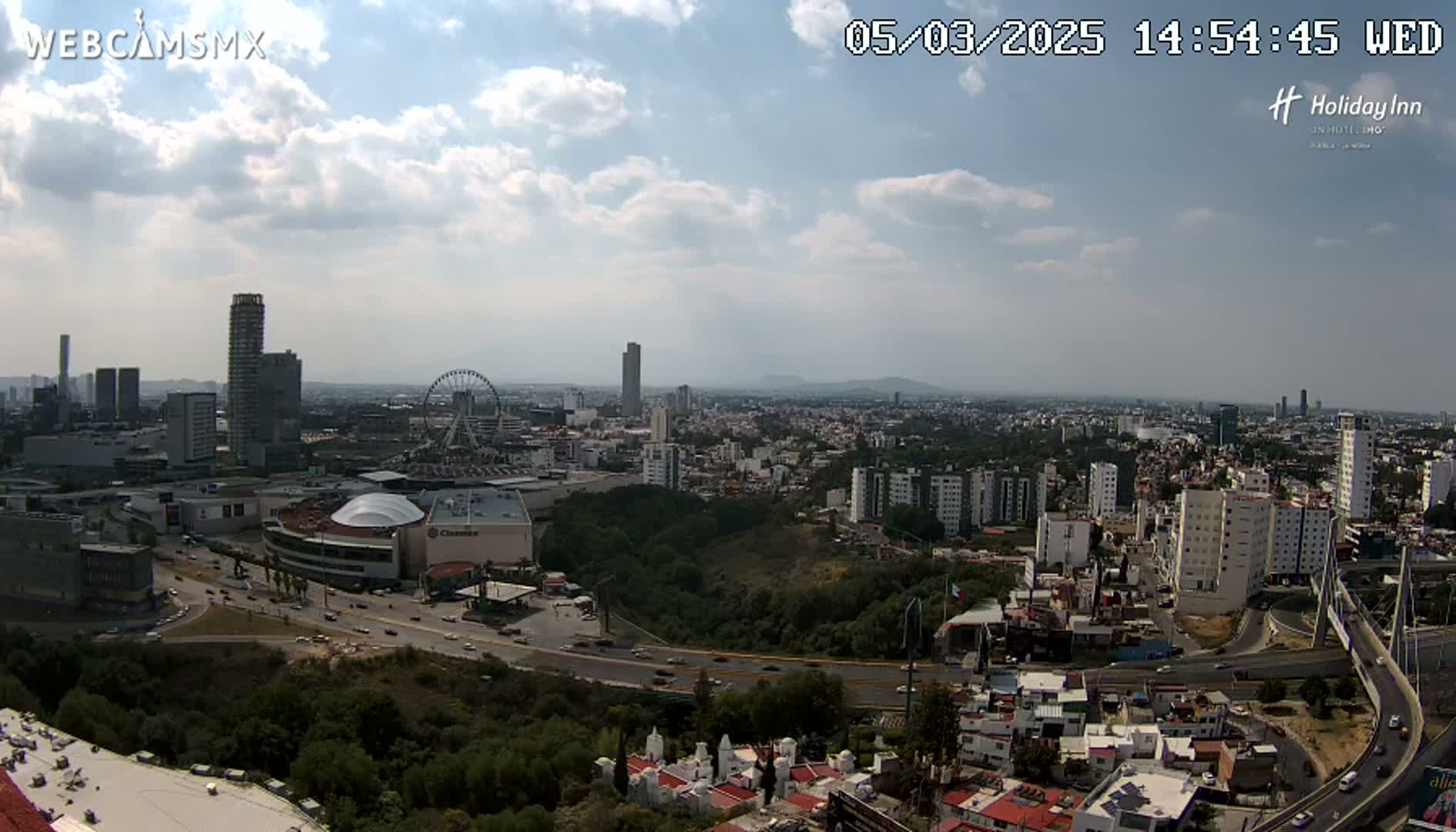 A partly cloudy daytime view of a sprawling city, featuring a Ferris wheel, several high-rise buildings, and a large shopping mall, with roads and surrounding green spaces visible.