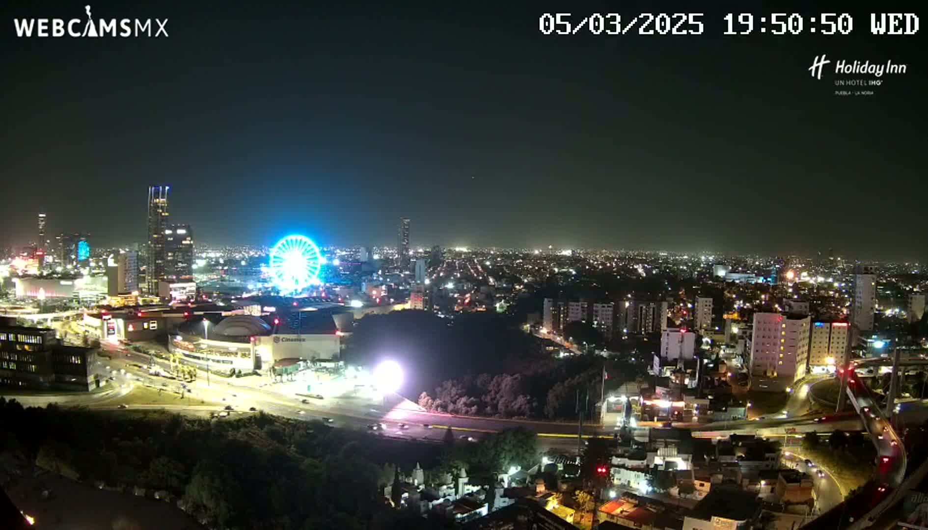 A nighttime, high-angle view of a city illuminated with lights, featuring a Ferris wheel and a large shopping mall, under a clear sky.