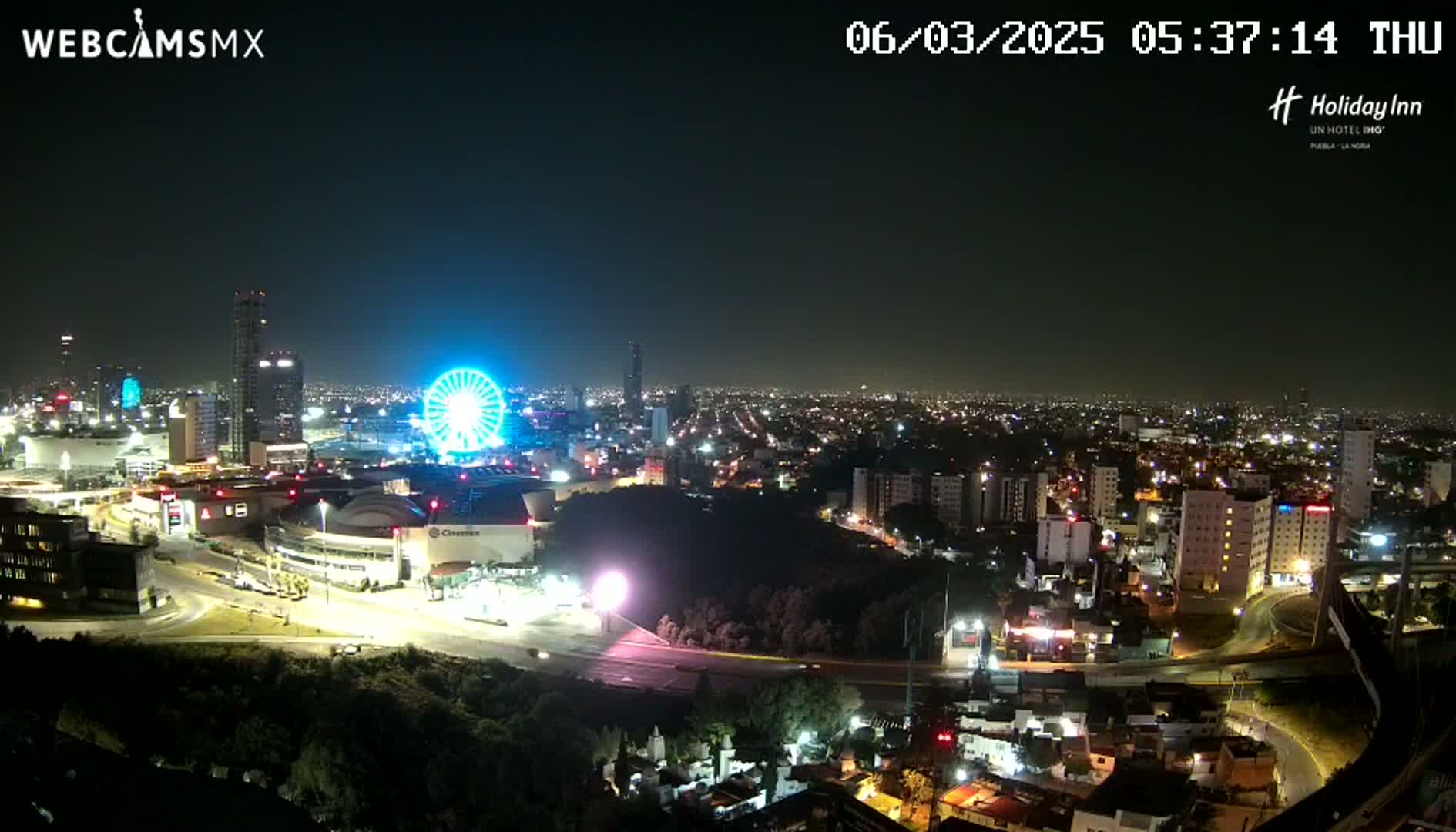 A nighttime view of a city illuminated with lights, featuring a Ferris wheel and several buildings, under a clear sky.