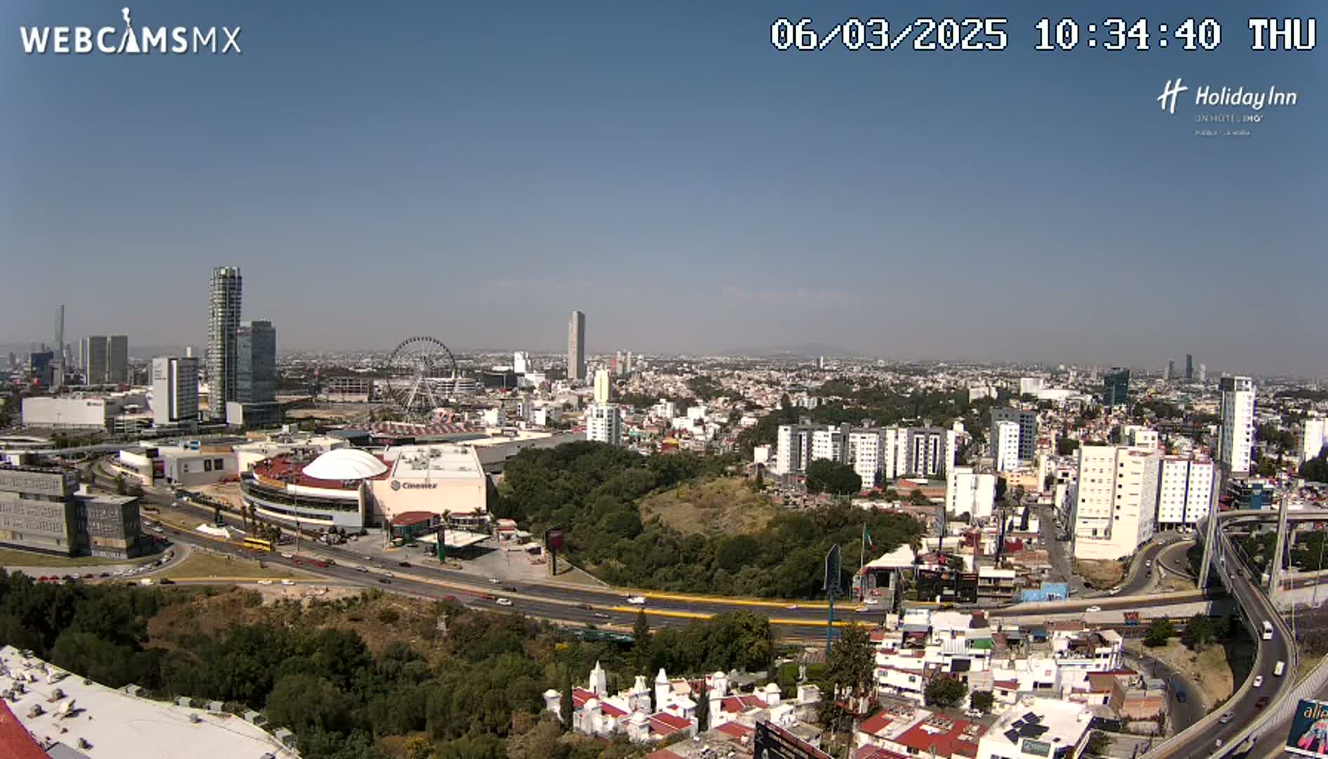 A sprawling cityscape under a clear blue sky, featuring a Ferris wheel, several high-rise buildings, and a large shopping mall complex next to a highway.