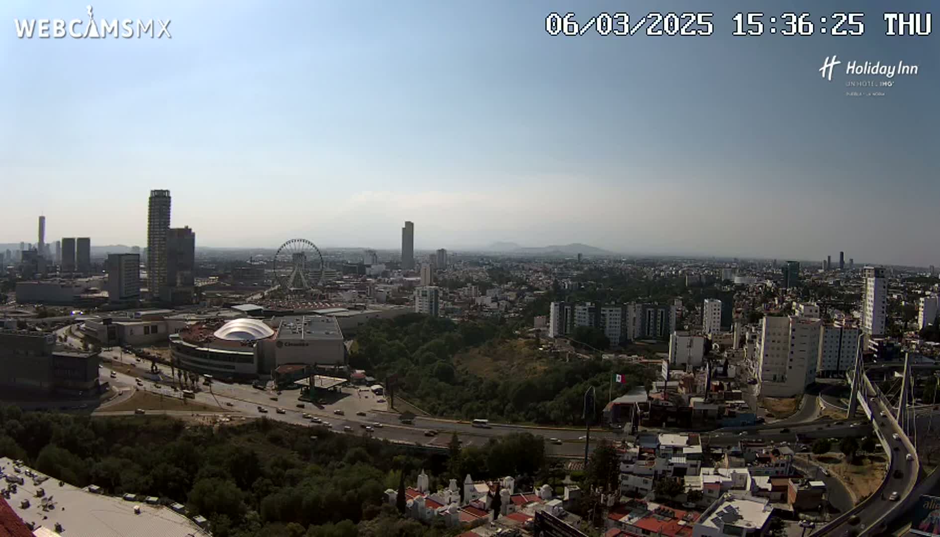 A high-angle, long shot on a clear, sunny day shows a sprawling city with high-rise buildings, a Ferris wheel, and a large shopping mall, all surrounded by a mix of residential areas and green spaces, with a highway weaving through the landscape.