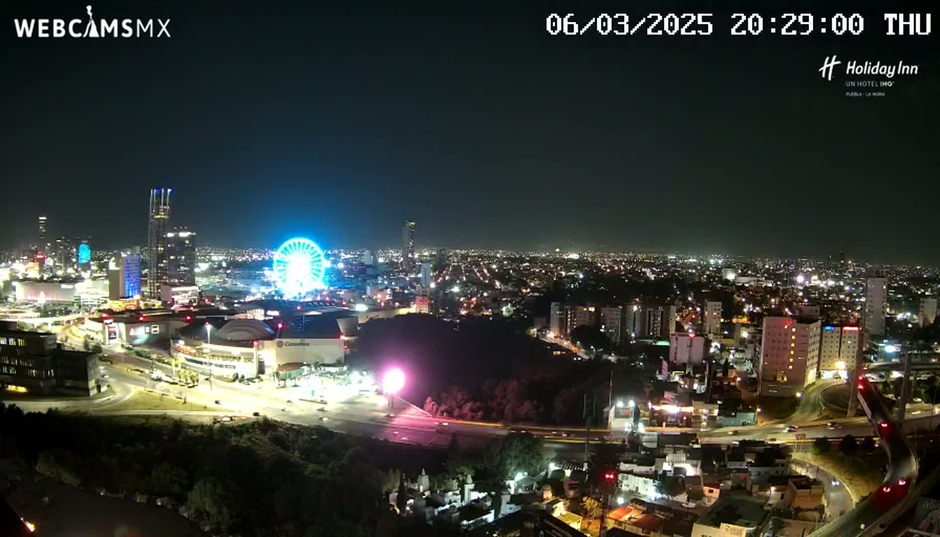 A nighttime, high-angle view of a city illuminated with lights, featuring a Ferris wheel and various buildings, under a clear, dark sky.