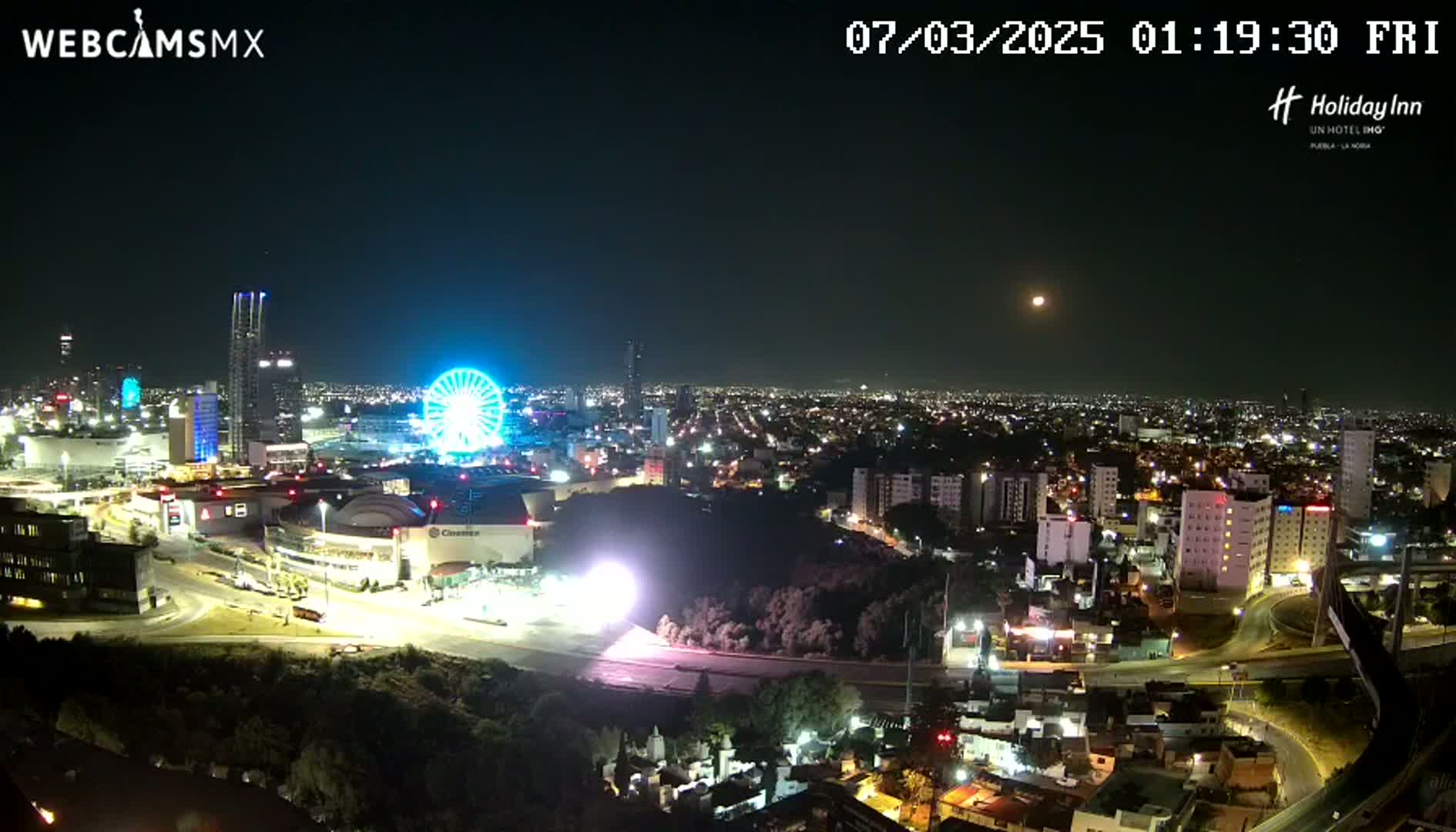 A nighttime aerial view of a city, illuminated by streetlights and featuring a brightly lit Ferris wheel, under a clear sky with a visible moon.