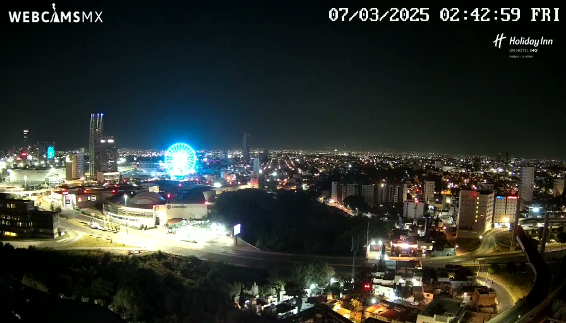 A nighttime aerial view of a city with a brightly lit Ferris wheel and numerous buildings, under a clear sky.