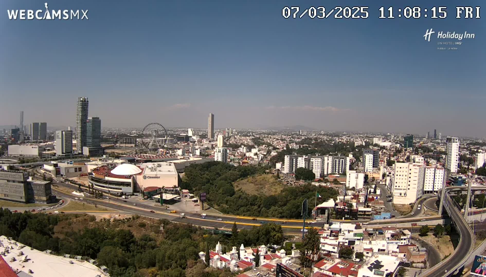 A sprawling cityscape under a clear, sunny sky shows a large shopping mall, Ferris wheel, and highway interchange amidst numerous buildings.