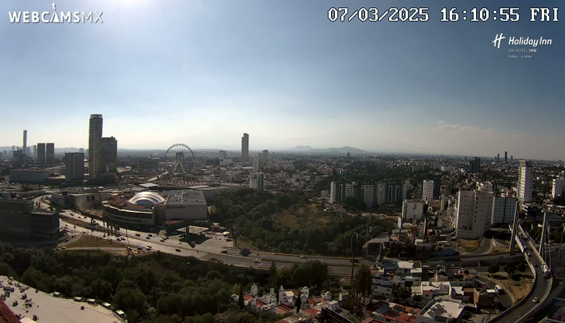 A sunny, high-angle view of a city with a Ferris wheel, a large shopping mall, and a bridge, surrounded by green hills and numerous buildings of varying heights.