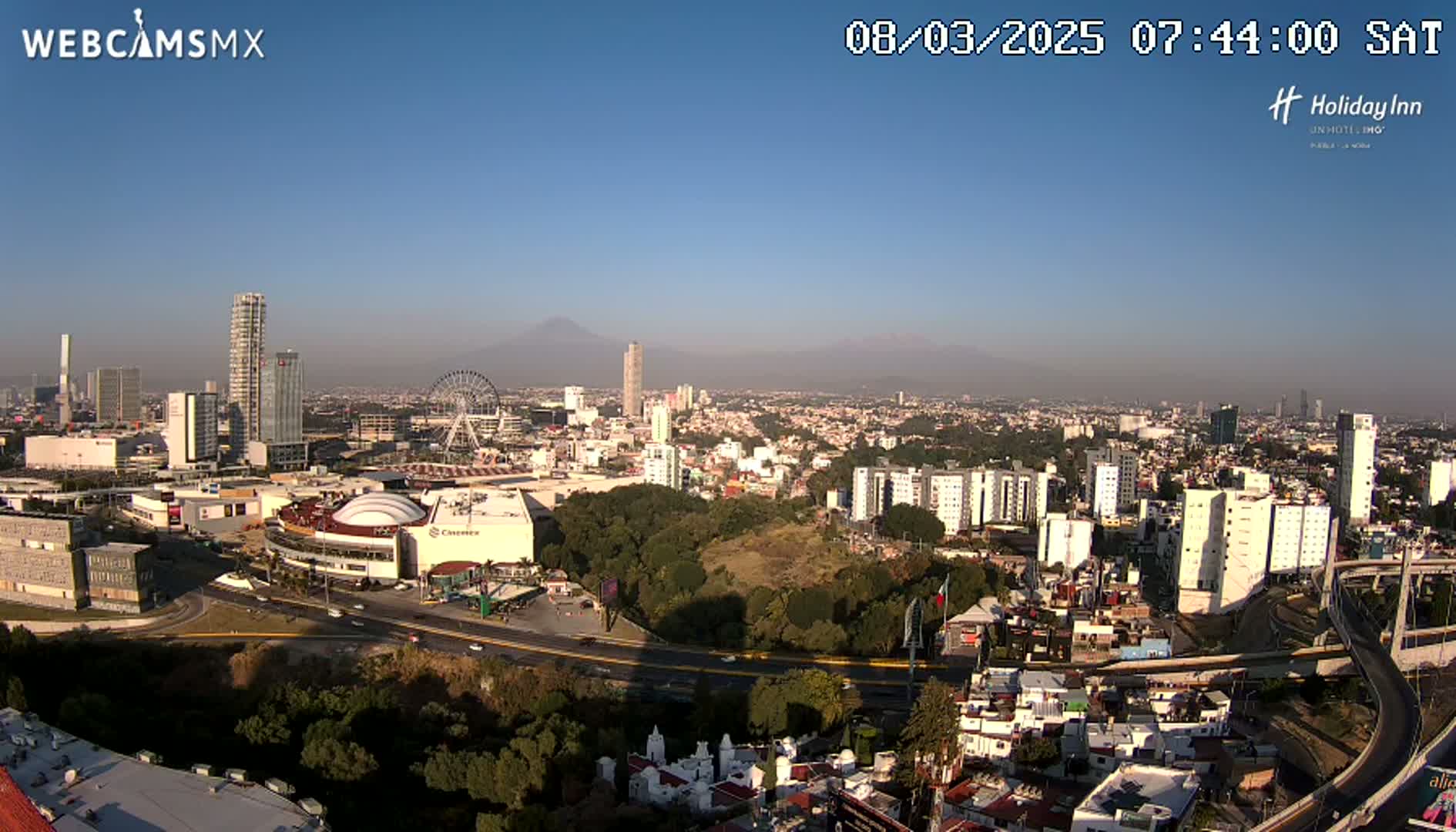 A high-angle, long shot on a clear, sunny day shows a sprawling city with numerous high-rise buildings, a Ferris wheel, and a large shopping mall, all situated in front of a mountain range in the distance.