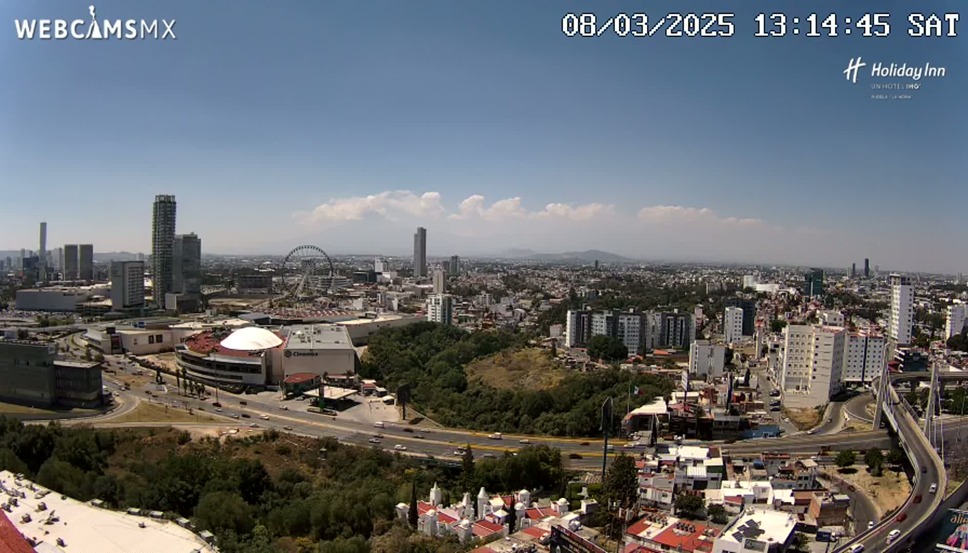A sprawling city landscape under a mostly clear, sunny sky, featuring a Ferris wheel, several high-rise buildings, and a large highway system curving through the scene.