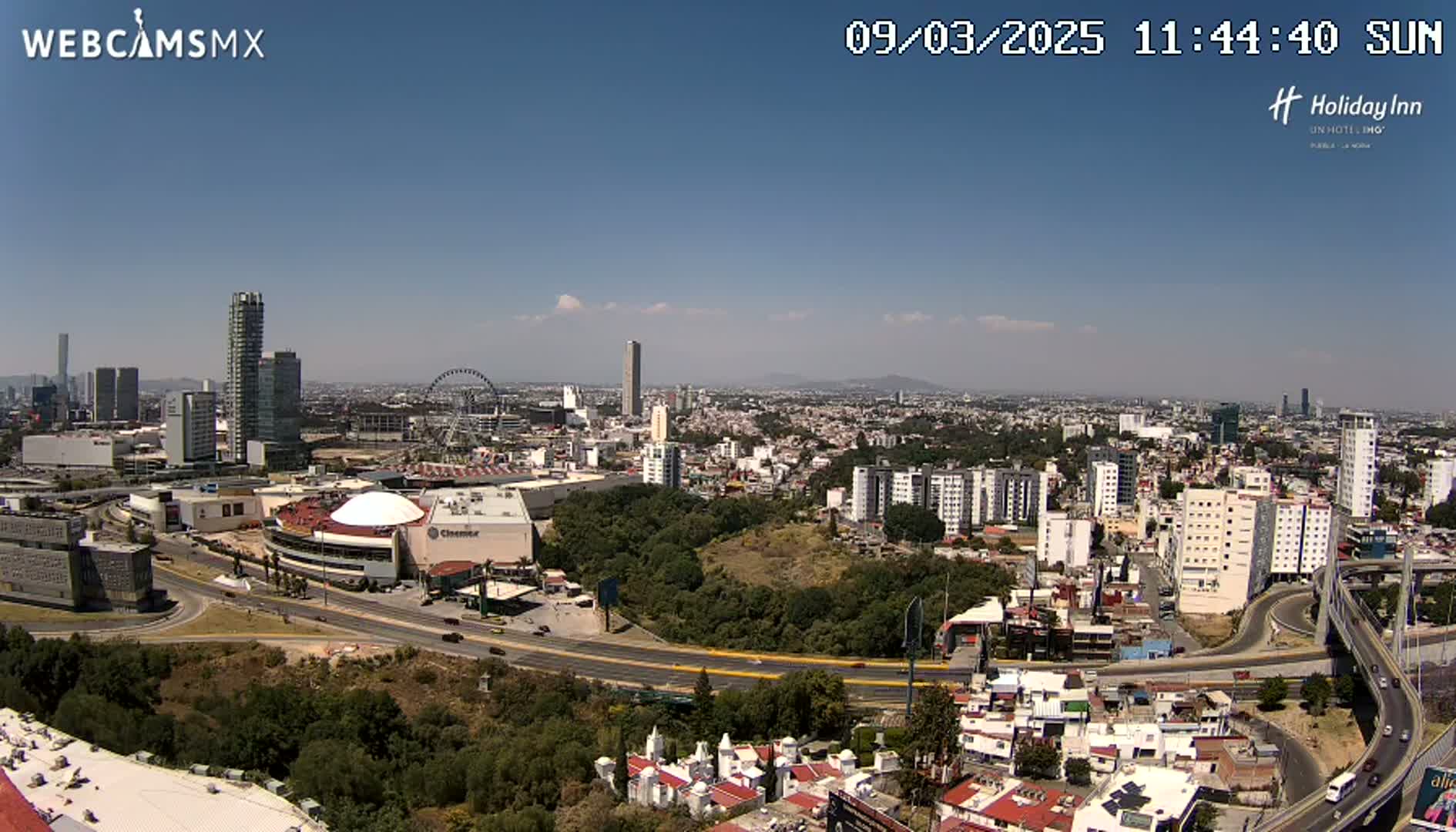 A sunny, high-angle view shows a sprawling city landscape with numerous buildings, roads, and a Ferris wheel, surrounded by green areas.
